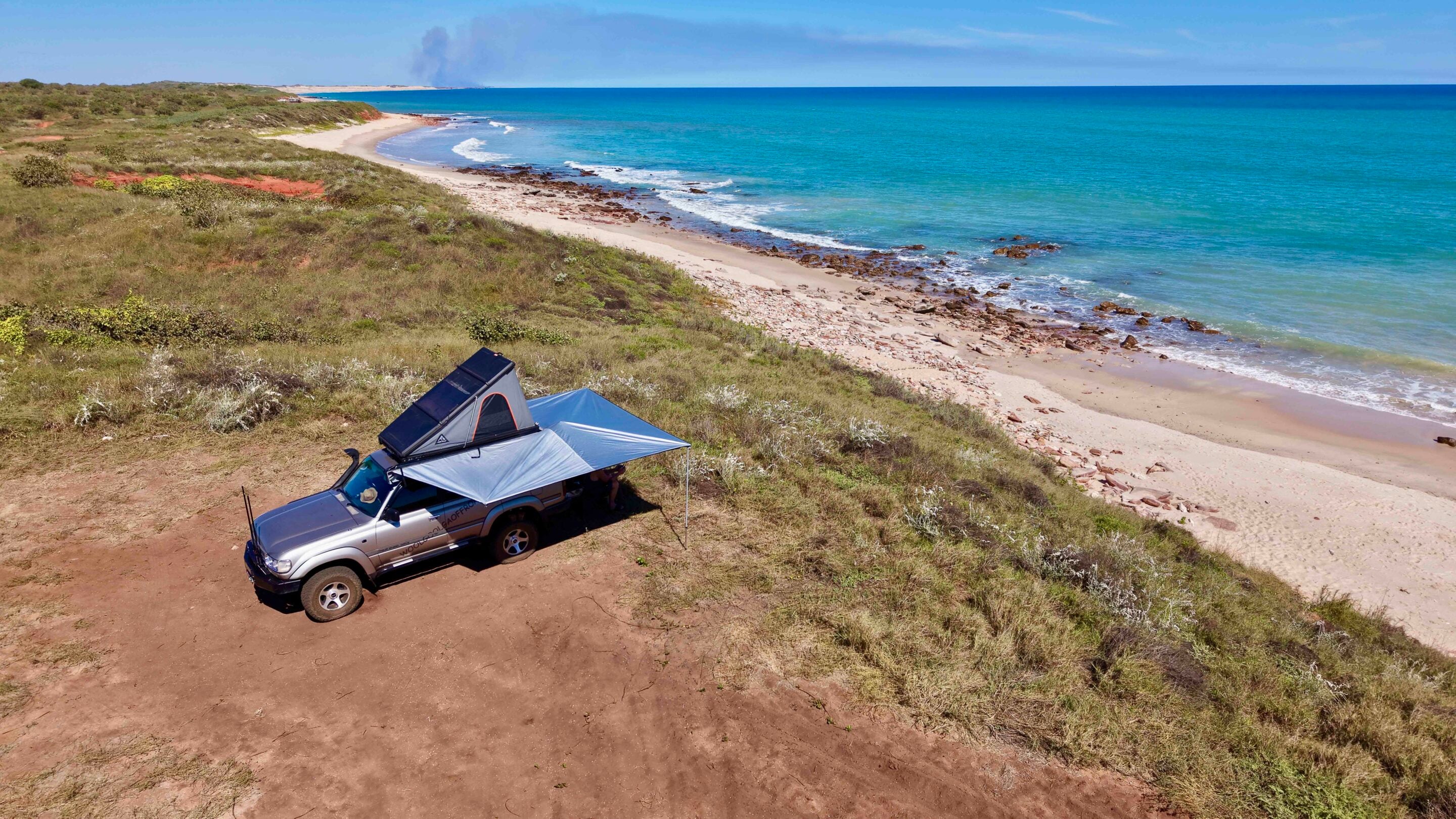SUV with rooftop tent parked on a sandy dune overlooking a rocky beach and blue ocean