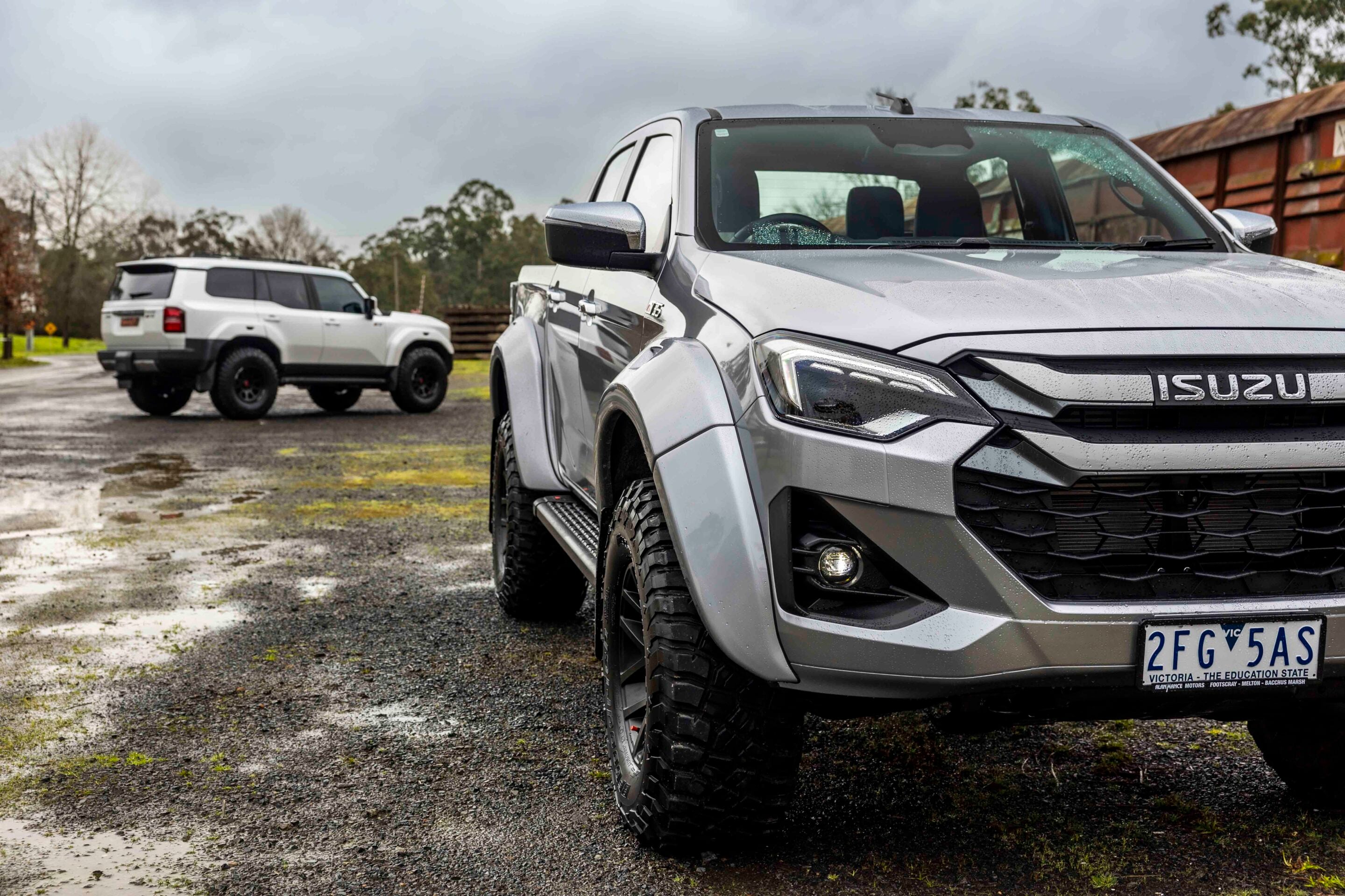 Close-up front-right view of a silver Isuzu pickup in a parking lot, with a white SUV in the background on a wet, muddy surface under a cloudy sky.