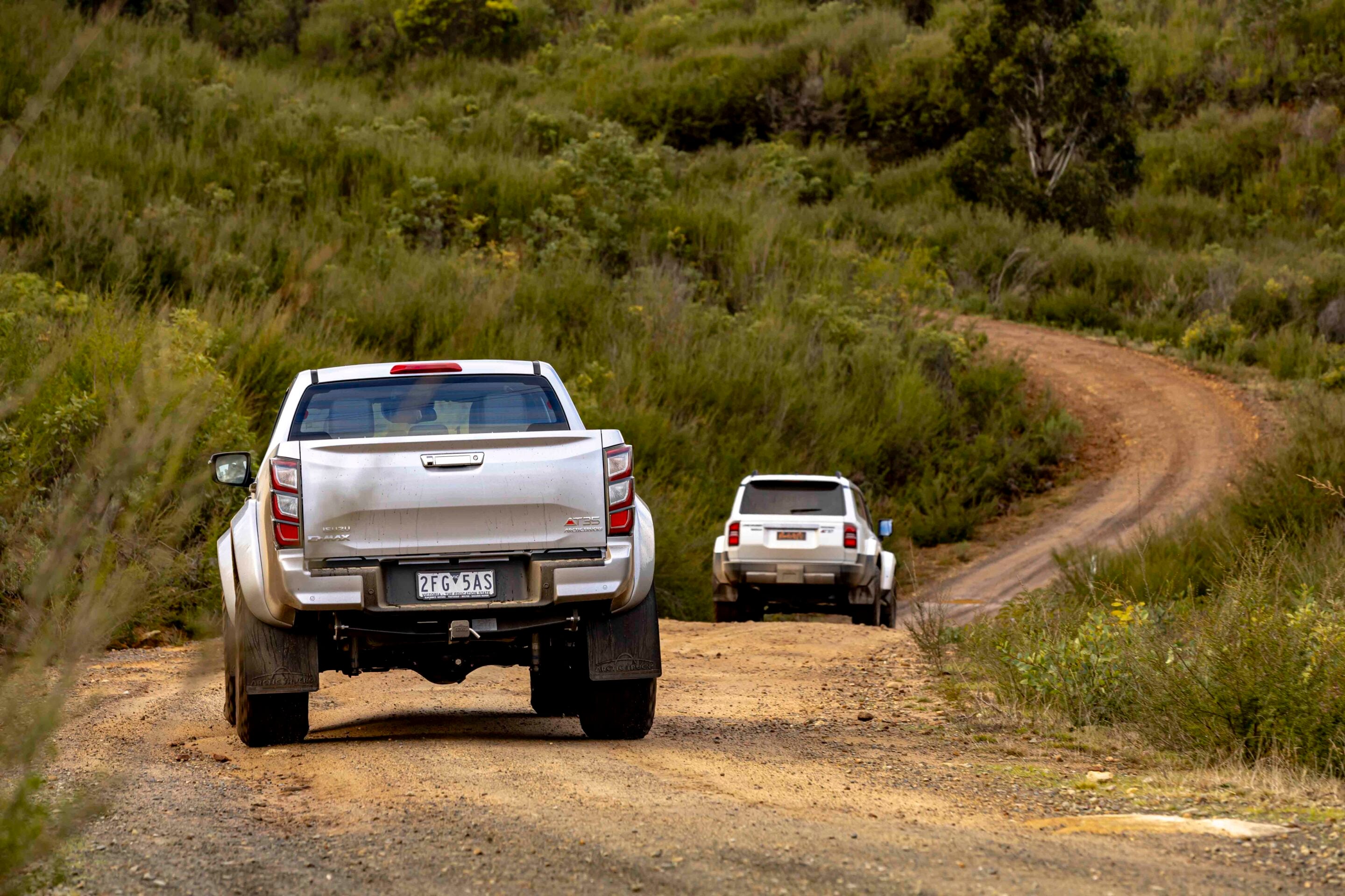 Three white pickup trucks driving on a dusty dirt road through a green, bushy hillside.