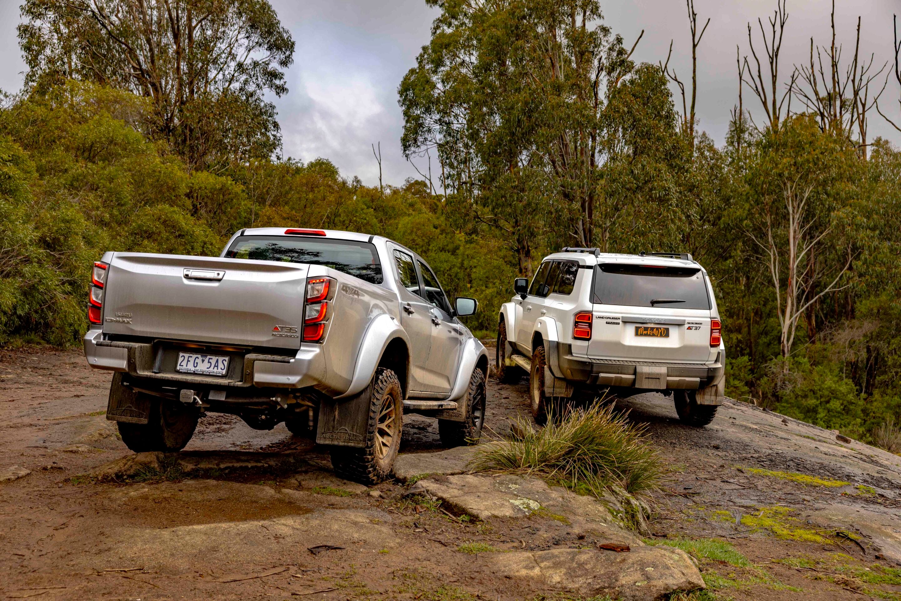 Two silver 4x4 pickup trucks parked on a muddy dirt trail in a forested area under an overcast sky.