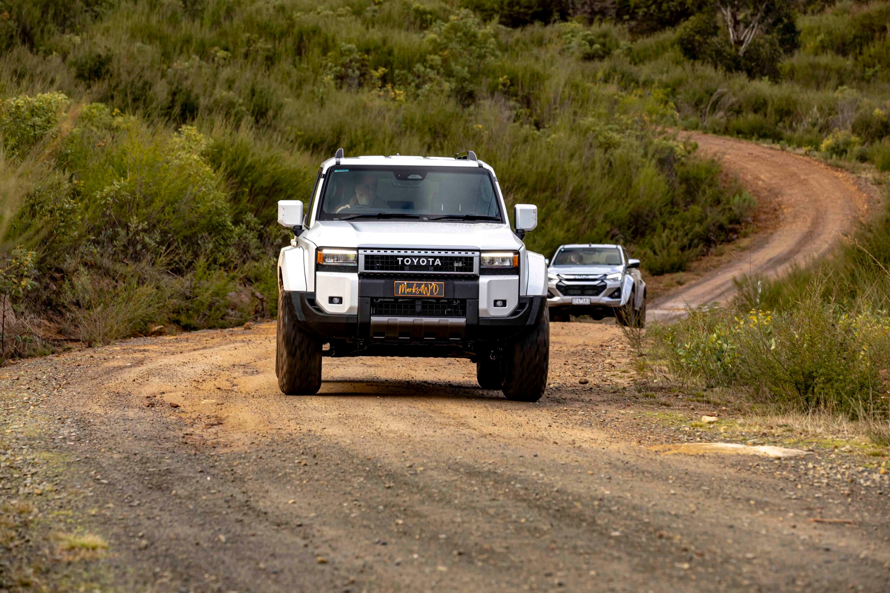 White Toyota SUV driving along a dusty off-road dirt road with a second vehicle in the background, surrounded by shrubs.