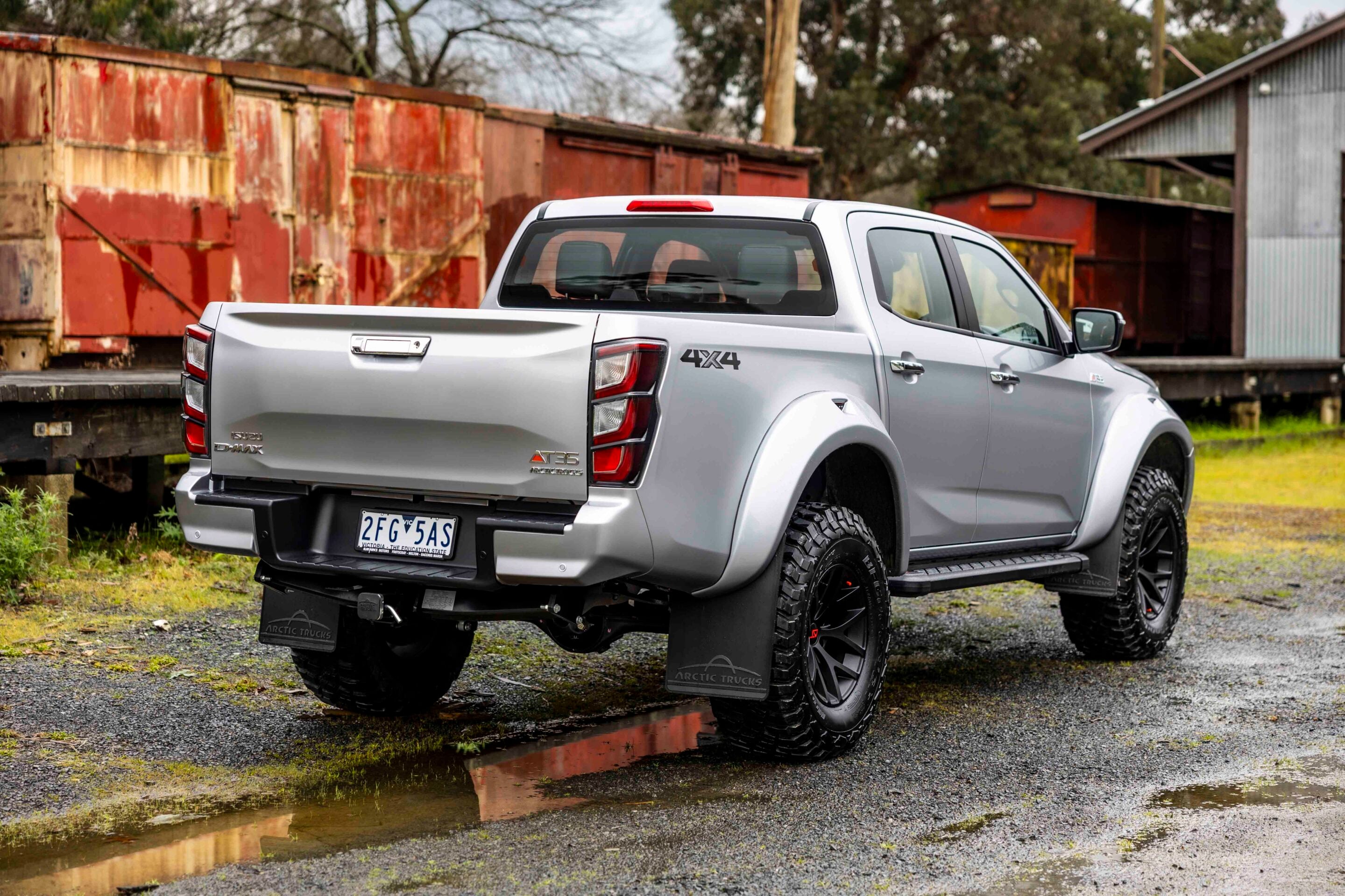 Silver four-by-four pickup parked on a gravel lot with rusted red shipping containers in the background and a muddy puddle nearby.