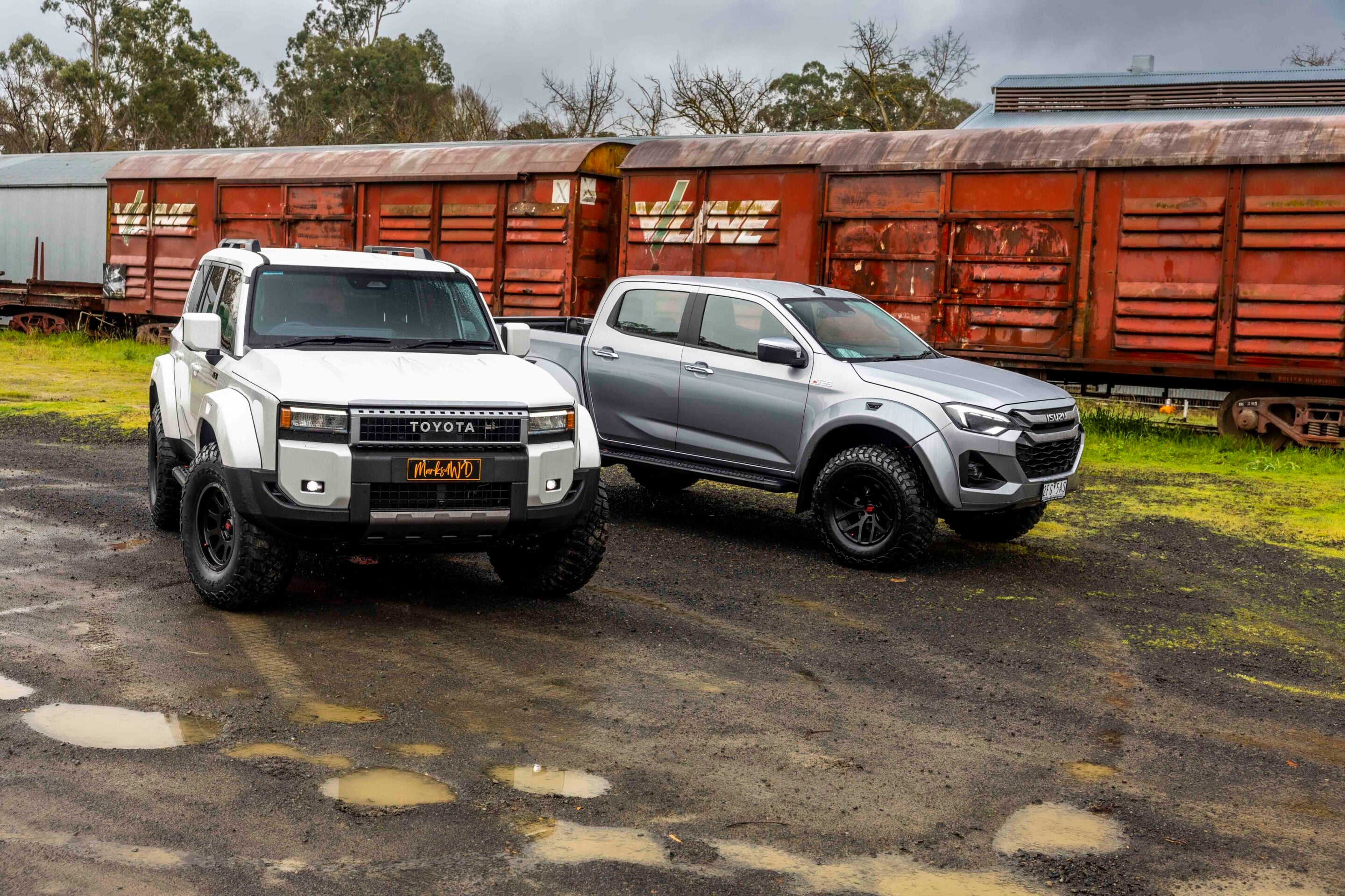 White Toyota pickup and silver Isuzu pickup parked in front of rusted red railcars on a muddy lot.