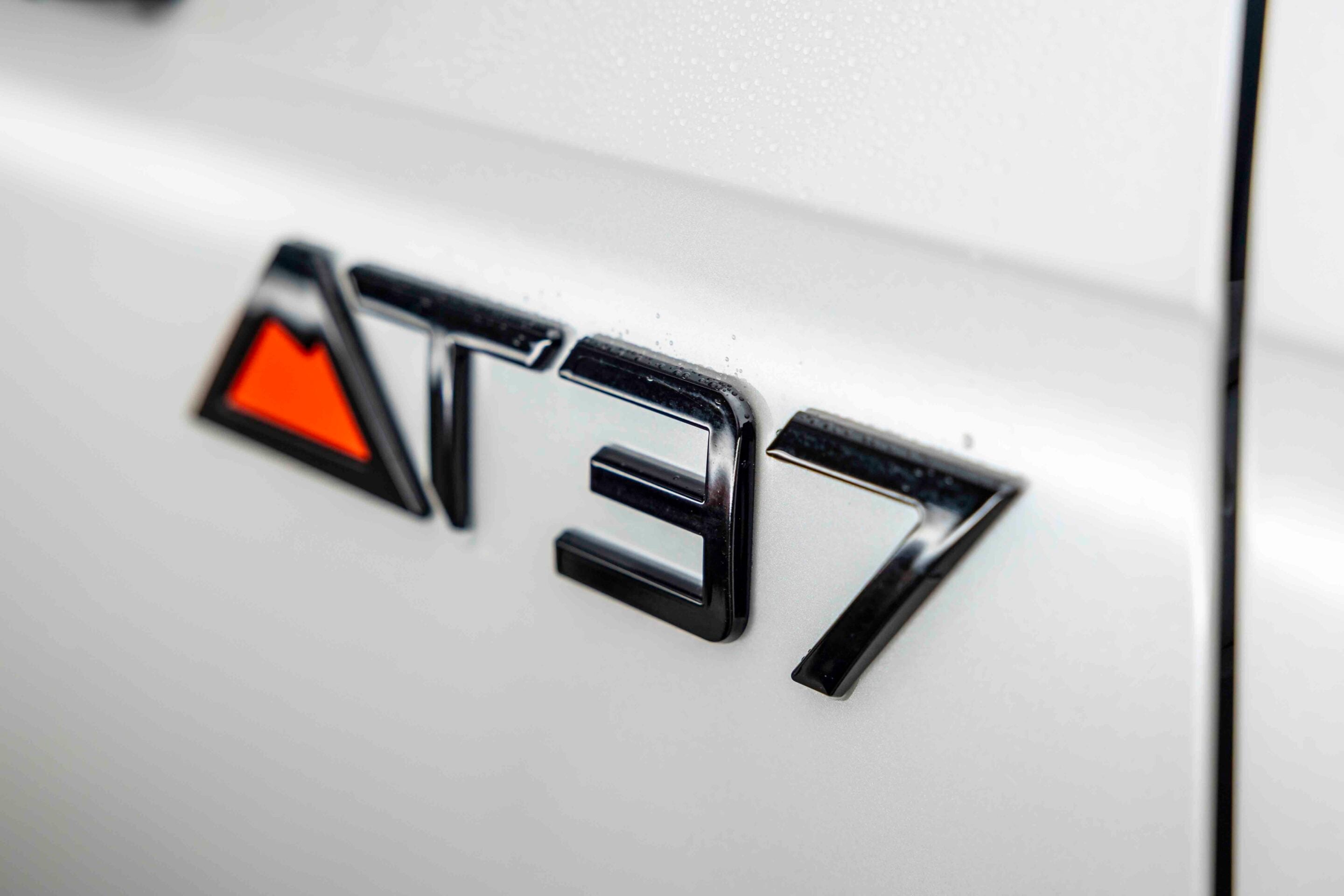 Close-up of a chrome car badge on a silver vehicle, with a partial orange triangular emblem visible to the left.