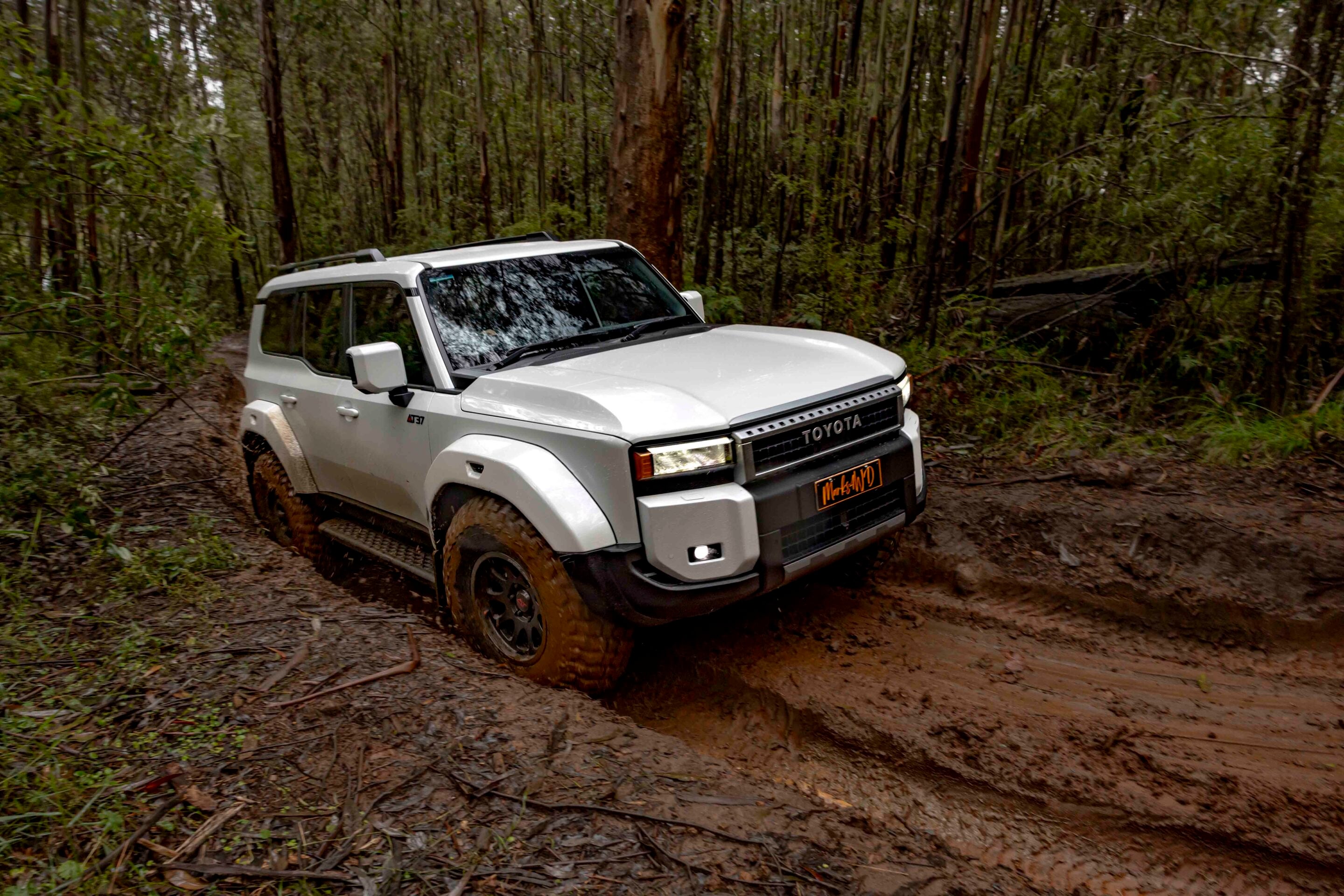 White Toyota SUV driving through a muddy forest trail, surrounded by dense trees.