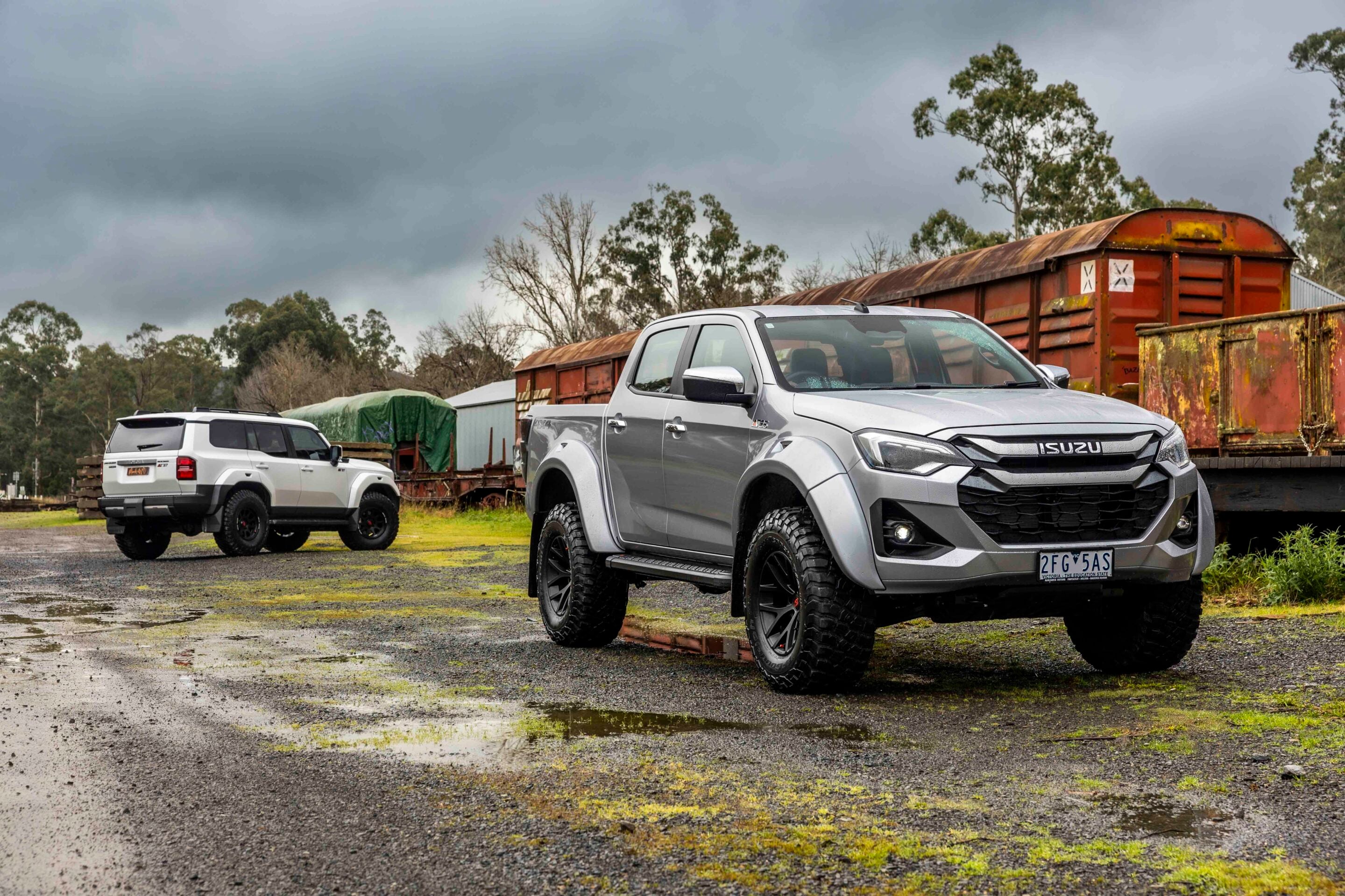 Gray Isuzu pickup in the foreground with a white SUV and rusted trailers in a muddy industrial lot under a dark cloudy sky.