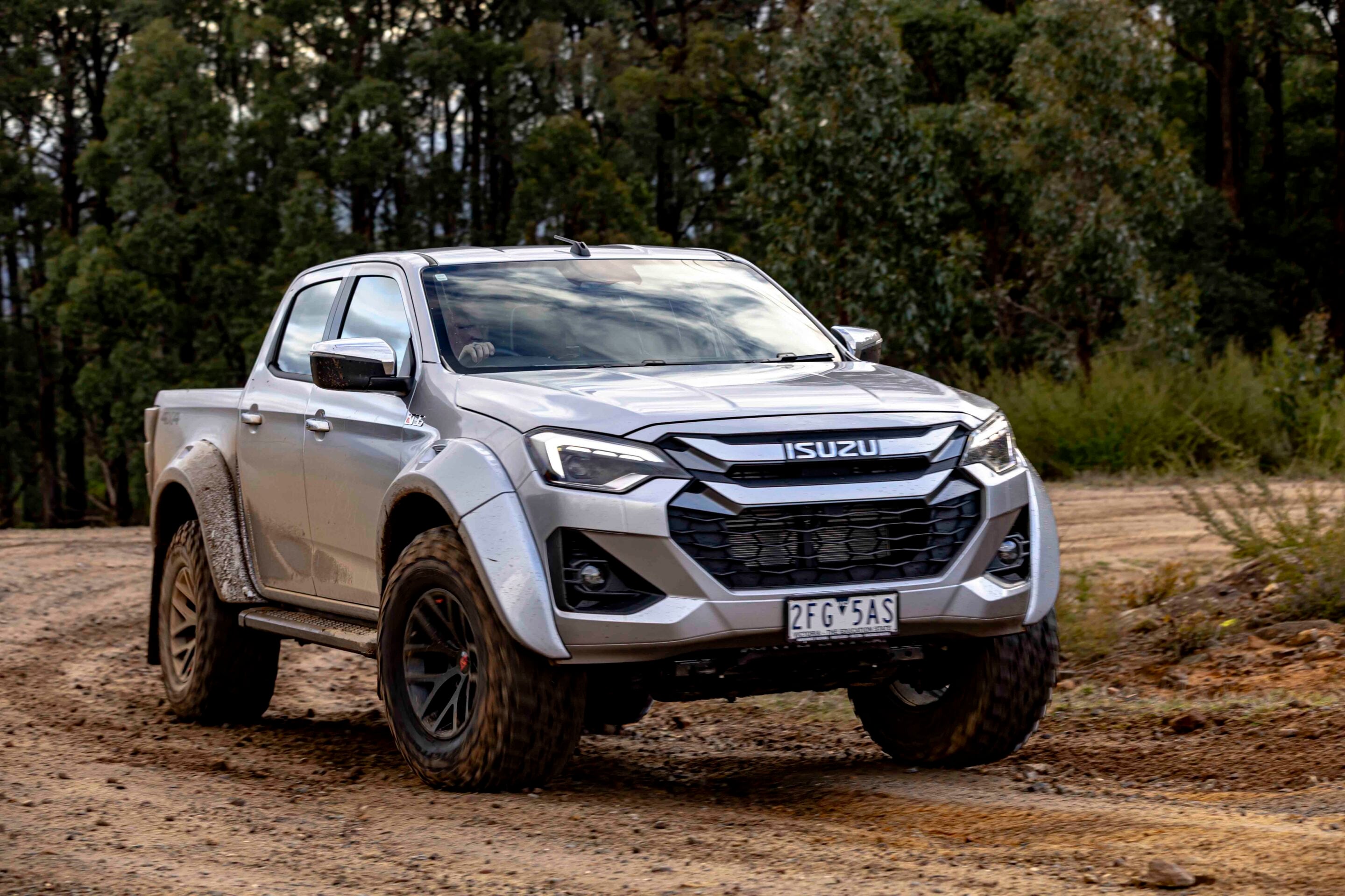 Front three-quarter view of a silver Isuzu pickup driving on a muddy dirt road with a wooded backdrop.