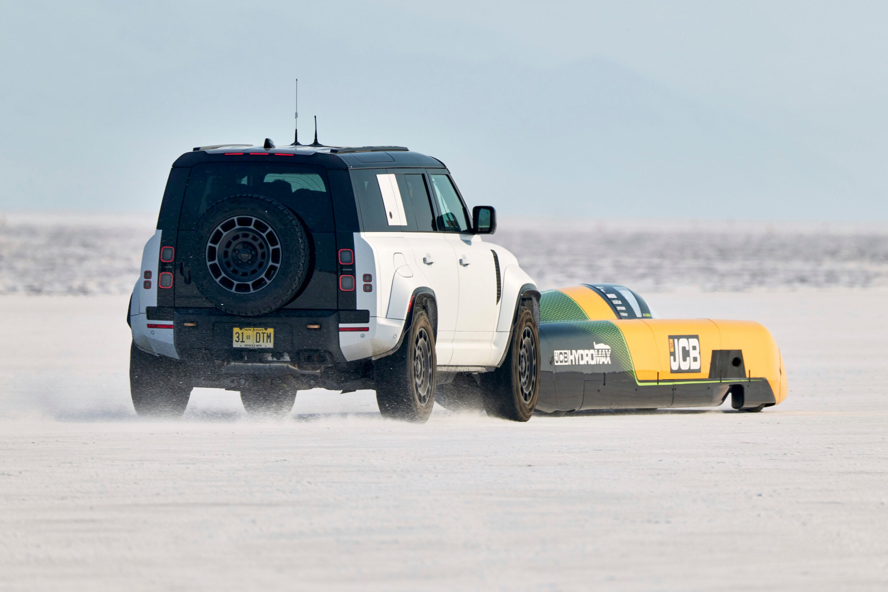 White and black SUV towing a bright yellow and orange JCB hydroplane on a snow/salt flat.