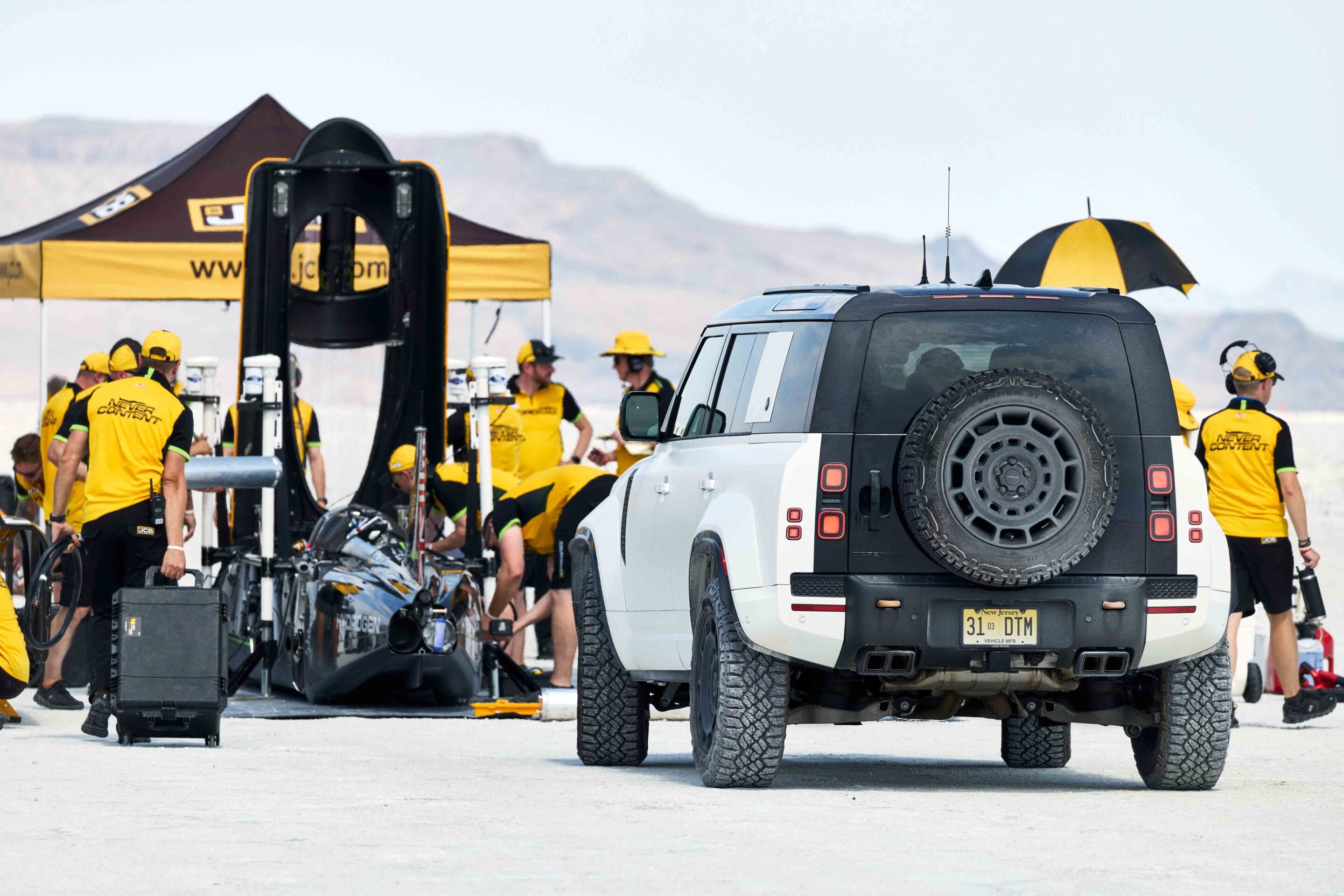Pit crew in yellow uniforms work around a white SUV with a mounted spare tire in a race pit area.