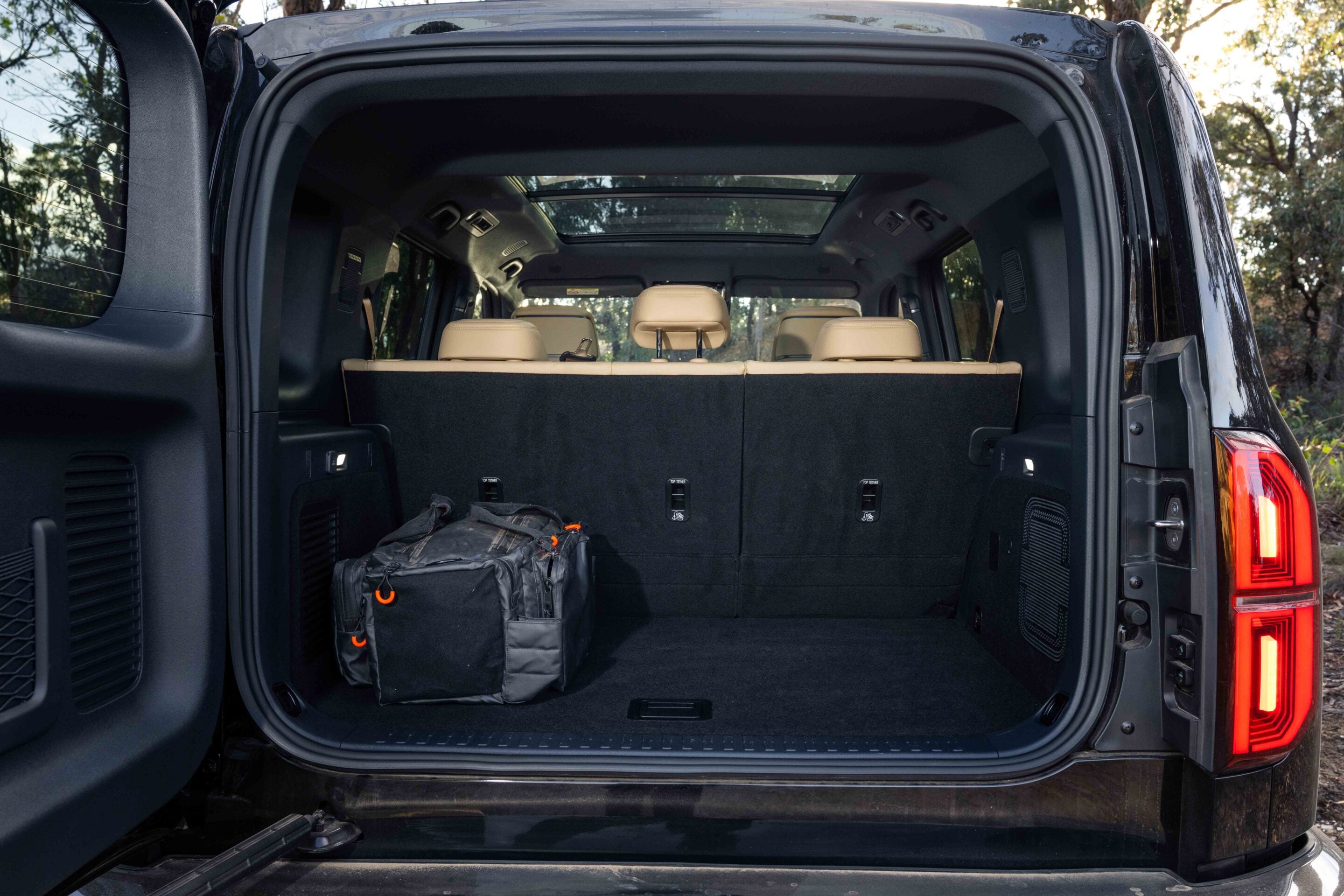 Open SUV trunk showing cargo area with two black bags and beige rear seats visible through the open tailgate, outdoor setting behind.