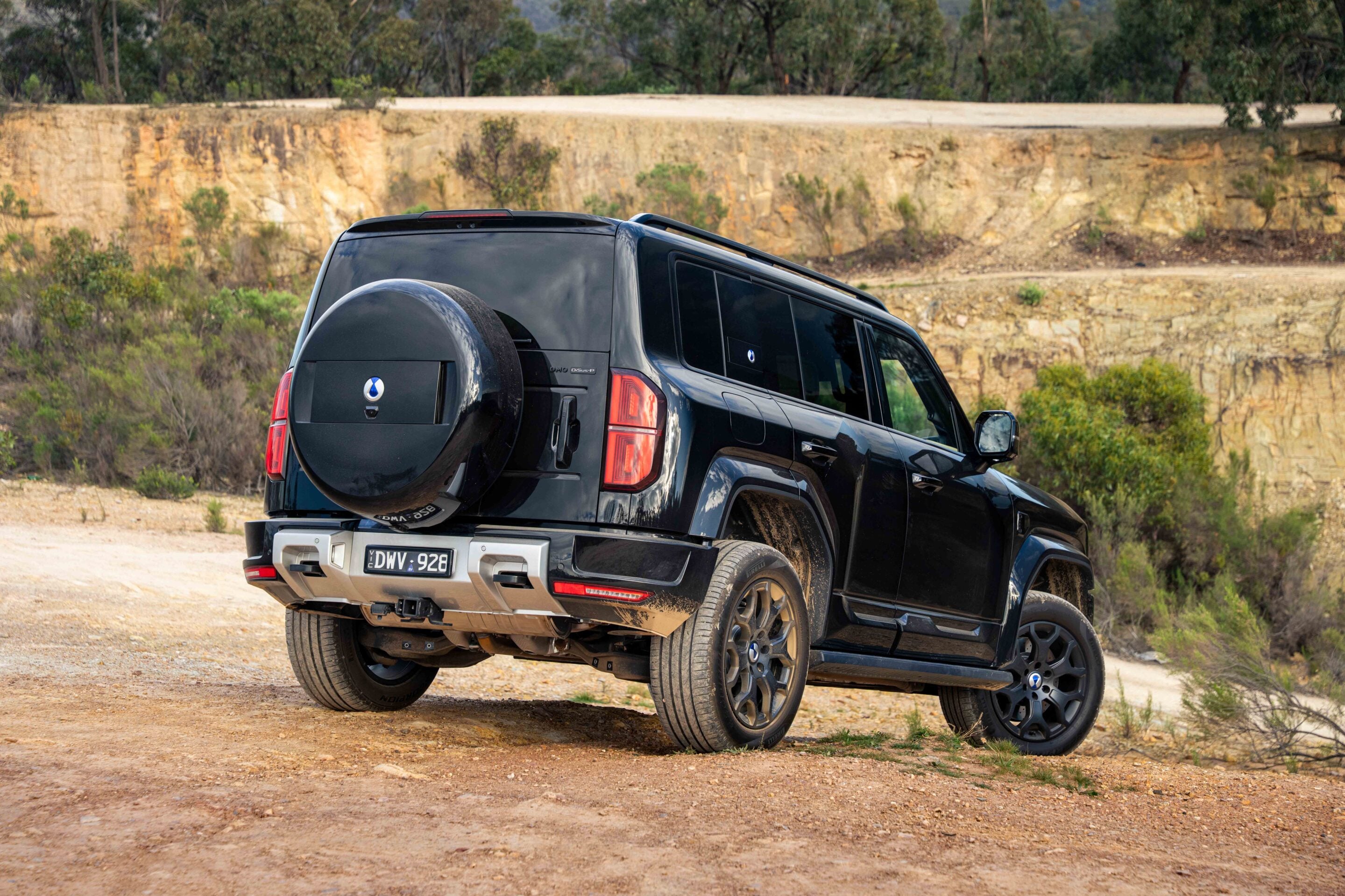 Black SUV (with spare tire mounted on back) parked on a dusty off-road trail beside a rocky hillside.