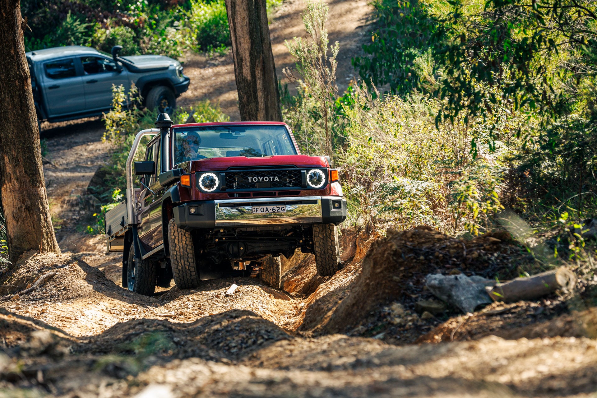 Red Toyota 4x4 navigating a rugged, rocky dirt trail through a sunlit forest with a silver pickup parked in the background