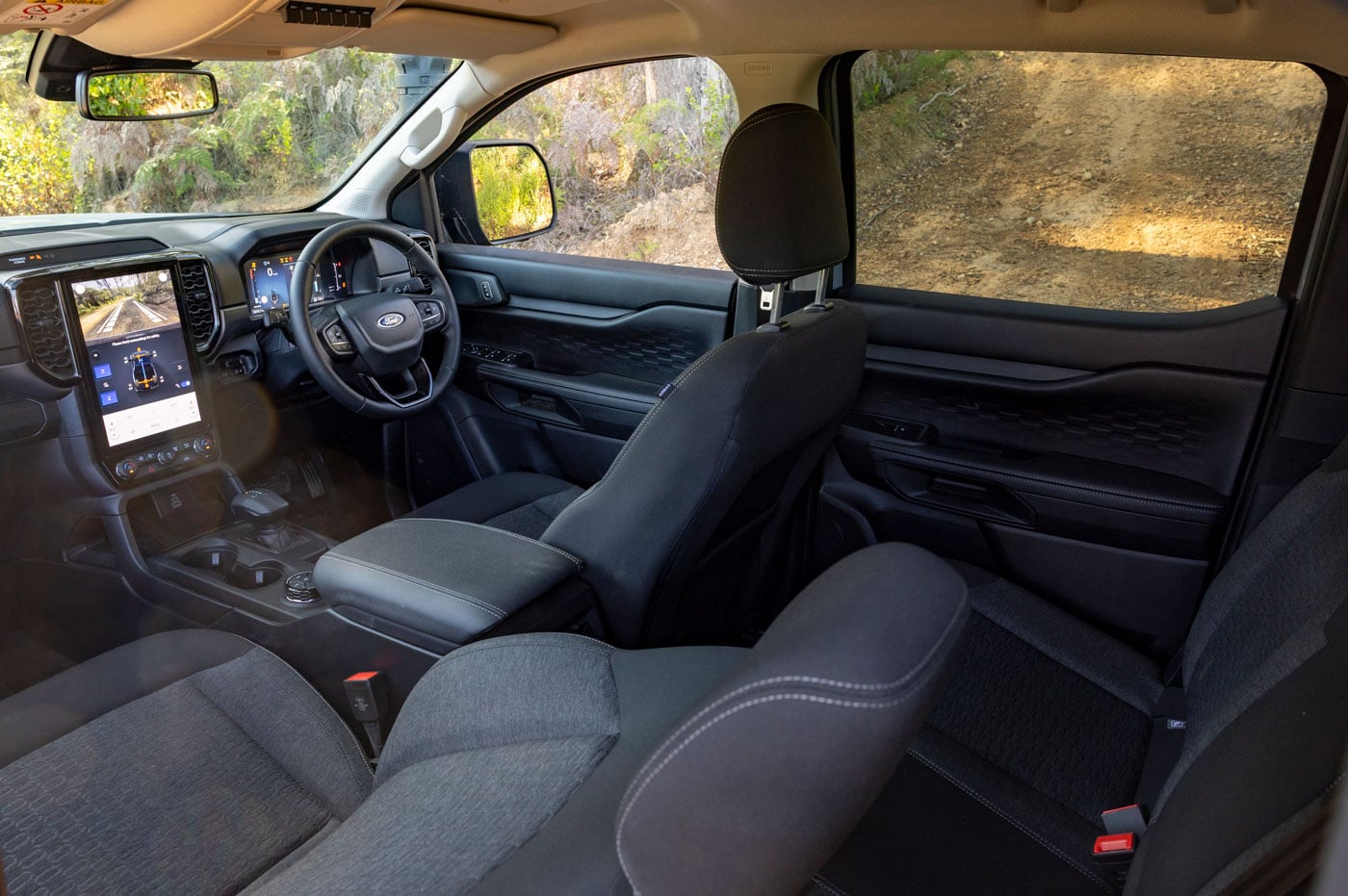 Interior of a modern pickup truck: driver and passenger seats, large central touchscreen, steering wheel, and gear shifter; view of rugged terrain outside through windows.