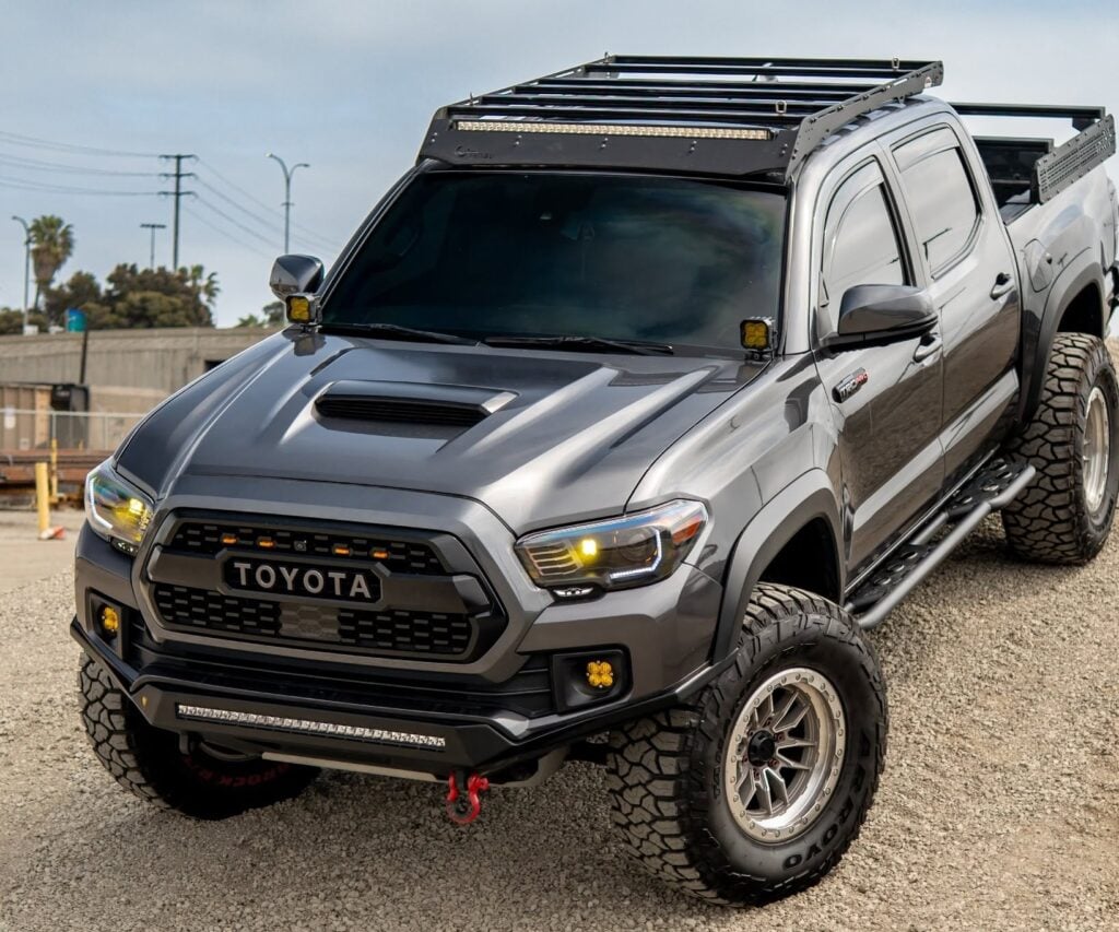 Grey Toyota pickup truck with a roof rack and off-road lights on a gravel surface, angled to show front and side.