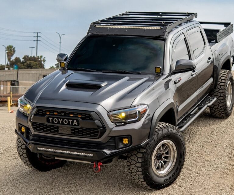 Grey Toyota pickup truck with a roof rack and off-road lights on a gravel surface, angled to show front and side.