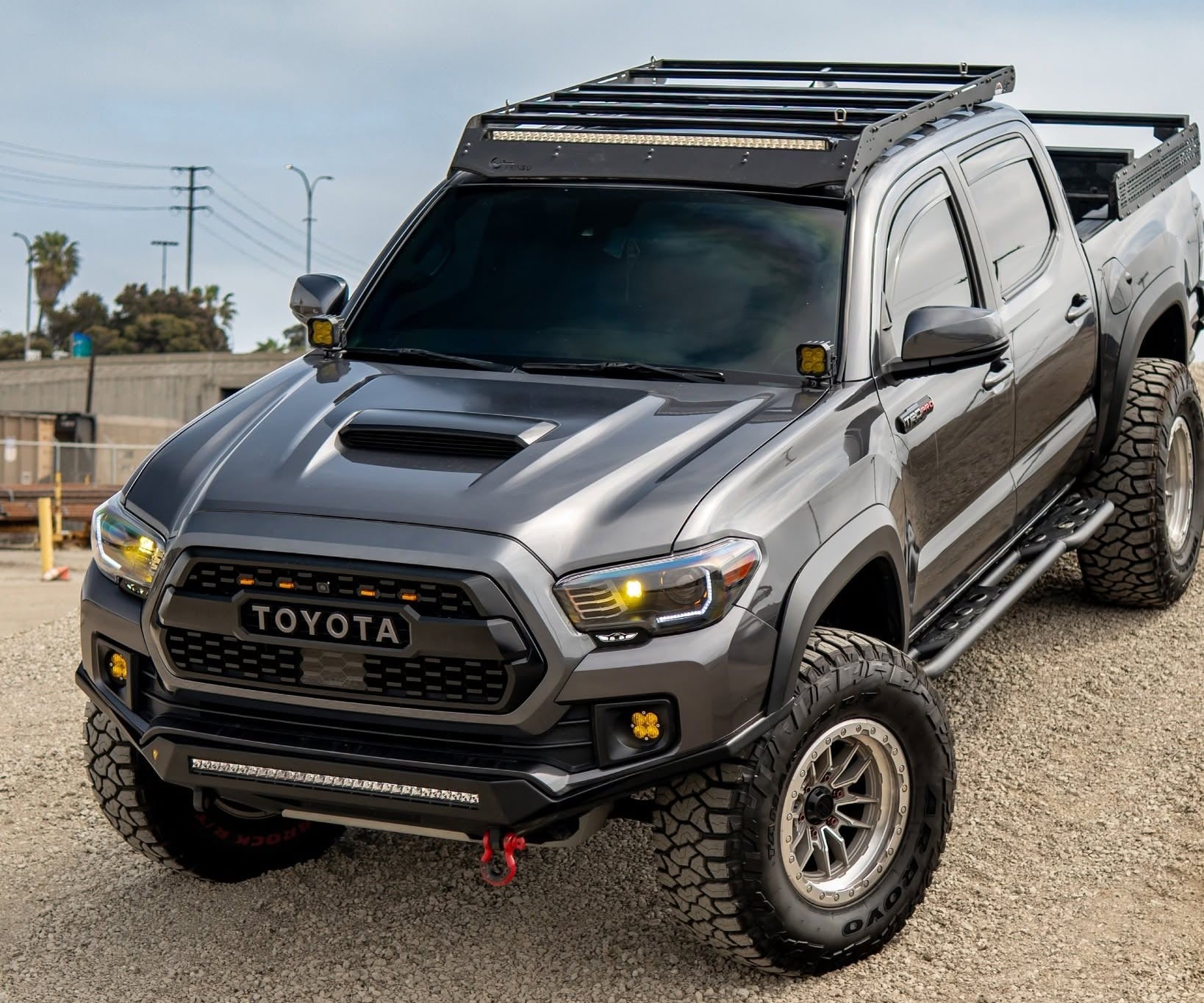 Grey Toyota pickup truck with a roof rack and off-road lights on a gravel surface, angled to show front and side.
