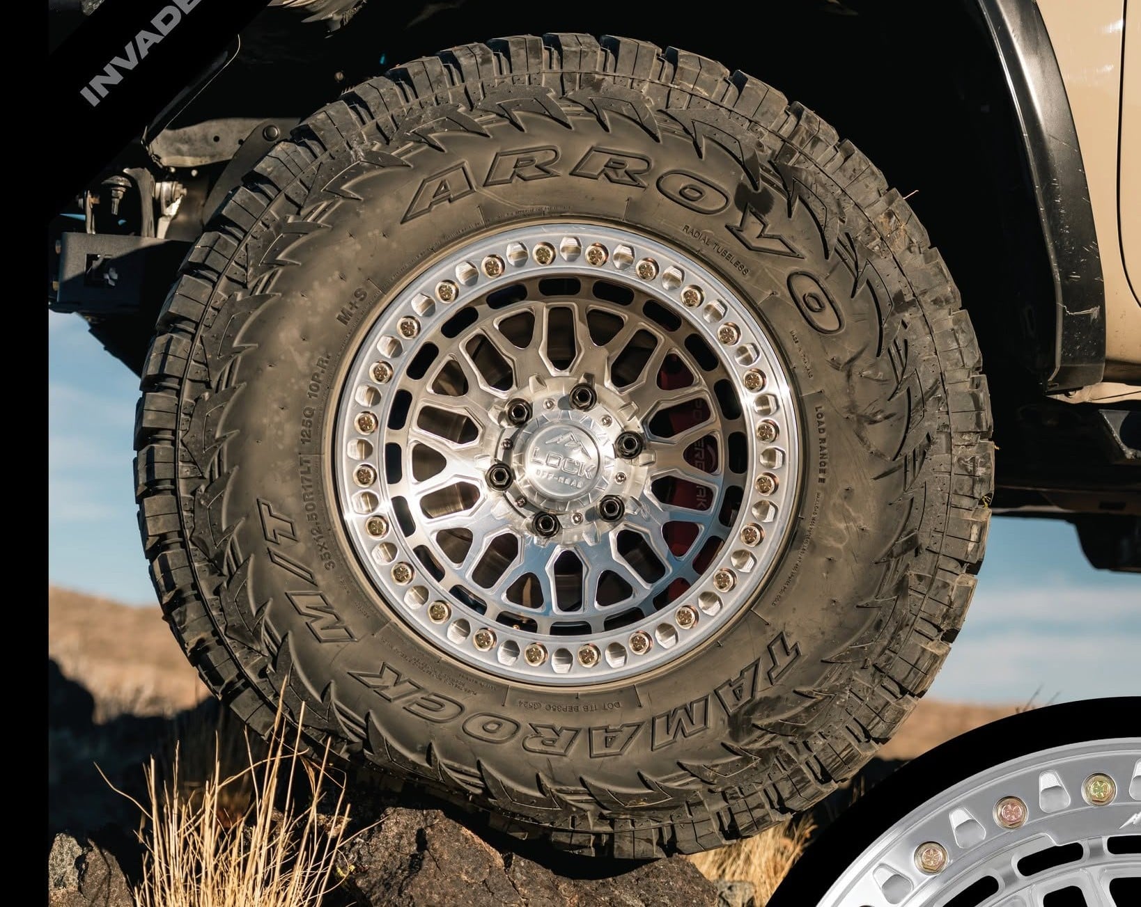 Close-up of a rugged off-road tire mounted on a chrome multi-spoke wheel, set against a rocky desert landscape.