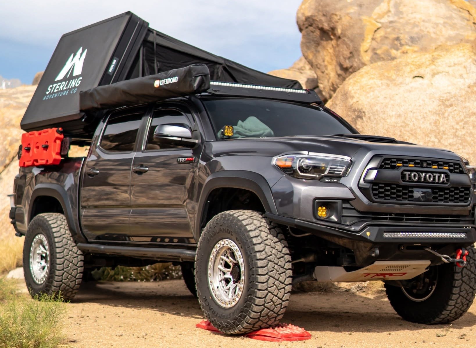 Black Toyota Tacoma with a roof-top tent and off-road gear parked in a rocky desert landscape; red jerry cans mounted on the side.