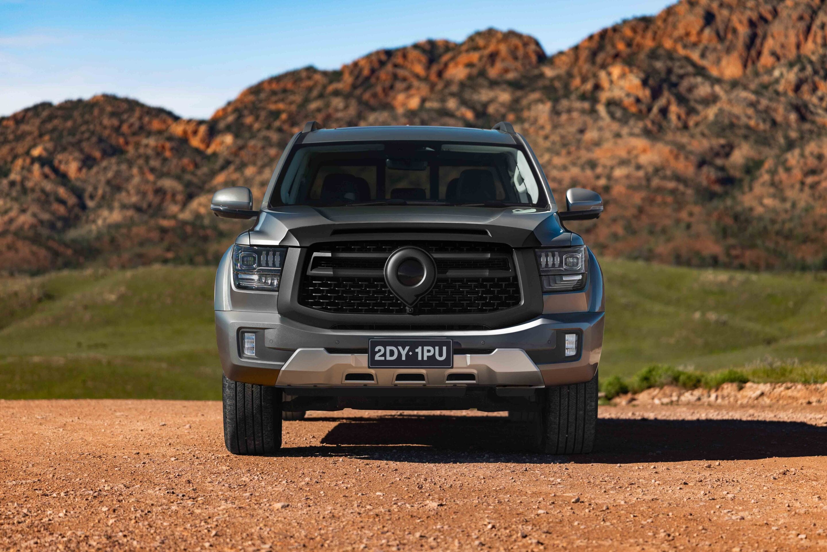 Front view of a gray pickup truck parked on a dirt road with rocky hills and a blue sky behind it.