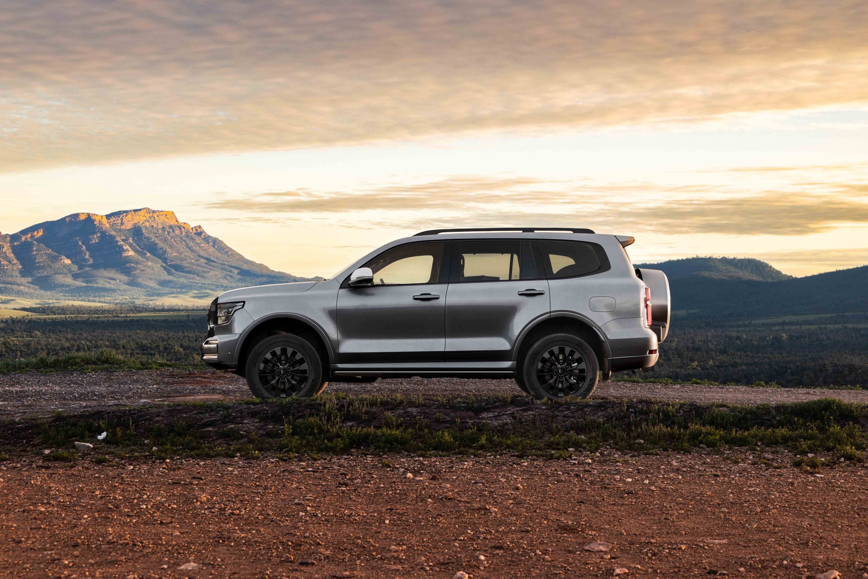 Silver SUV parked on a dirt road with rugged mountains at sunset in the background.