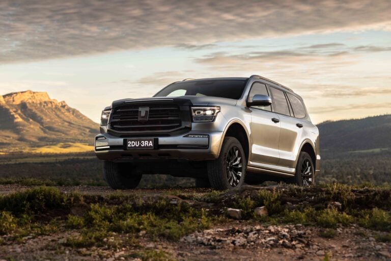 Silver SUV on a rocky dirt road with mountains in the background at sunset, front-left view showing grille and headlights.