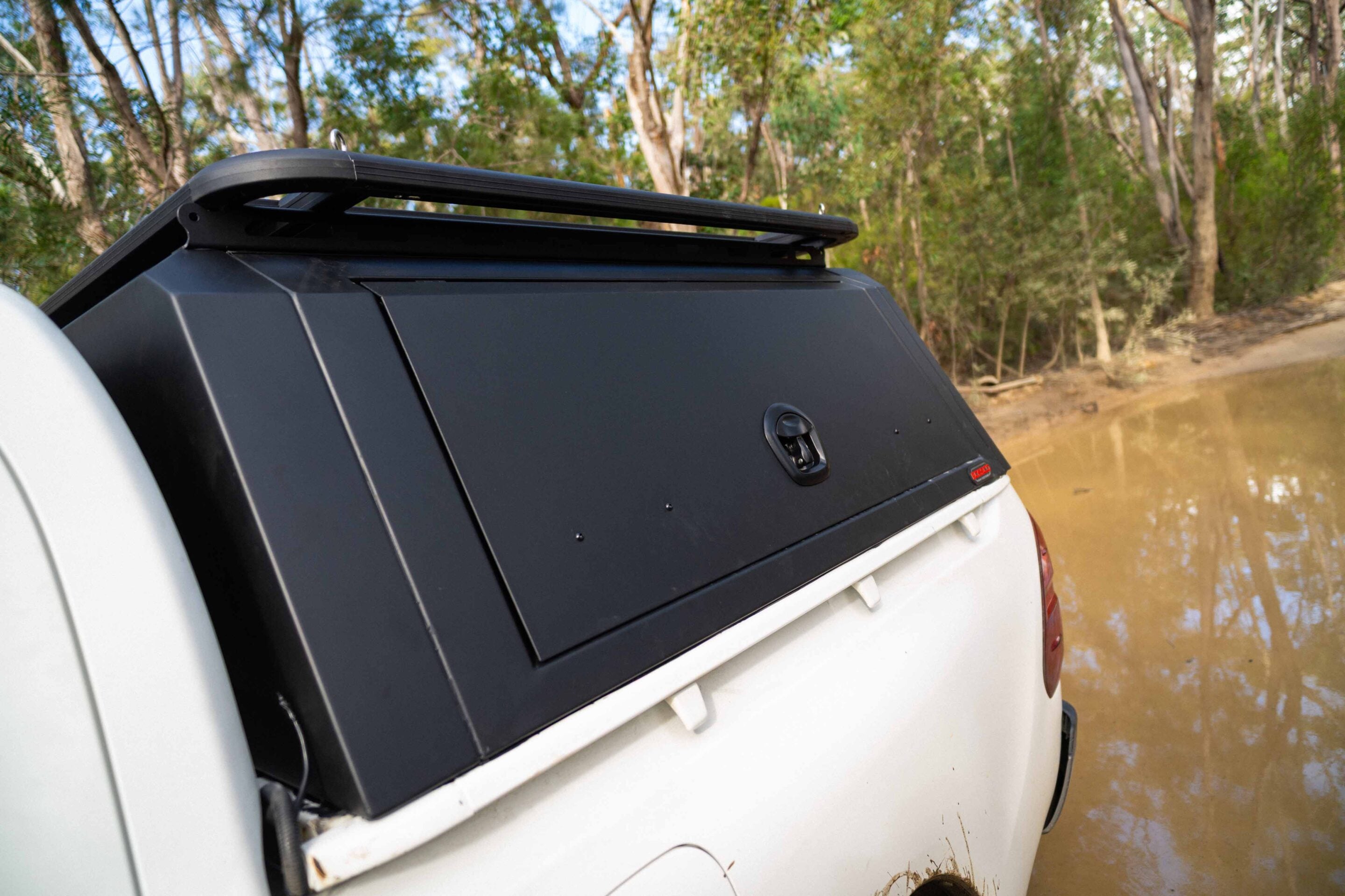 White SUV with a black roof rack parked by a muddy riverbank in a forested area.