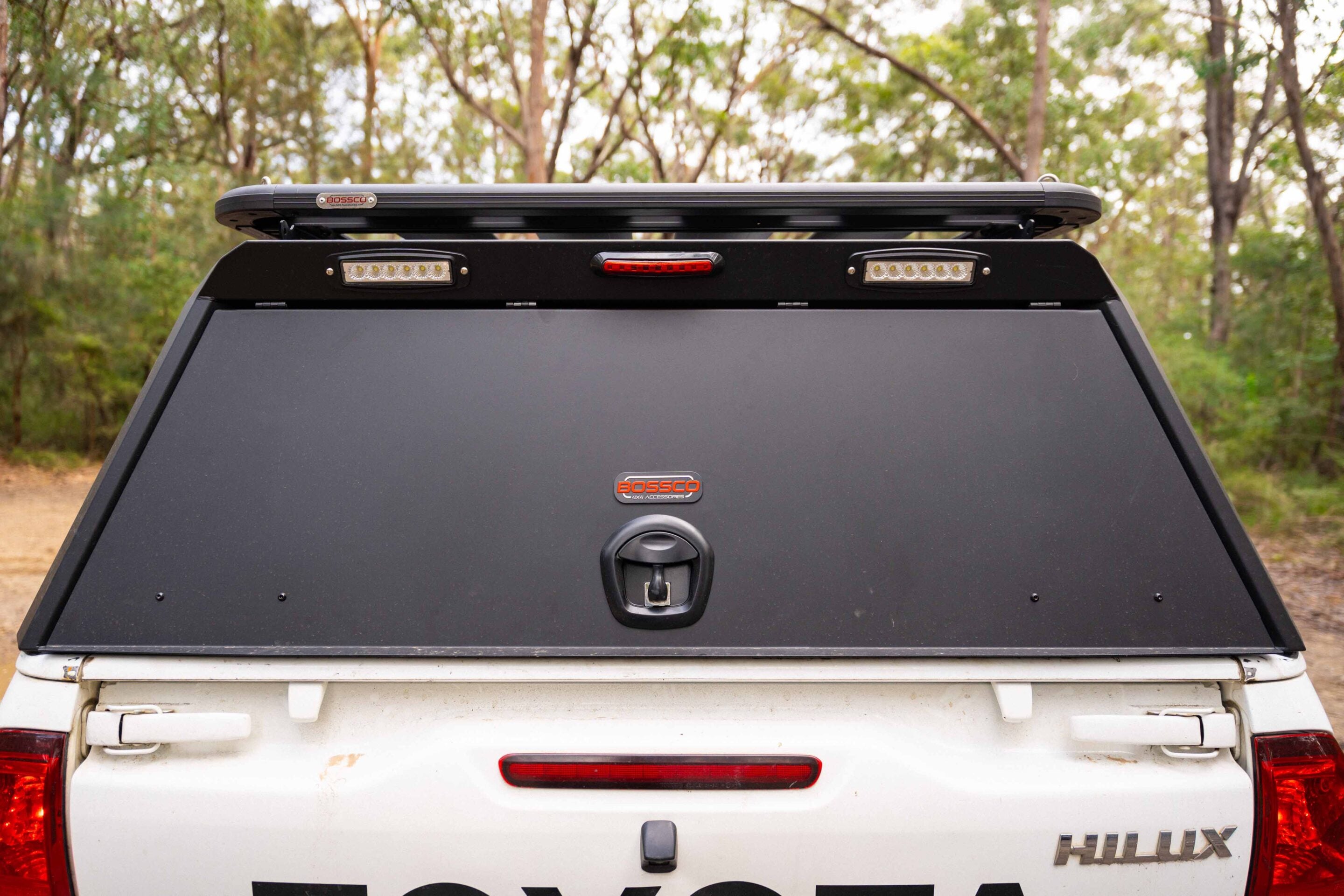 Rear view of a white Toyota Hilux with a black canopy, roof rack/line and dual LED lights on the rack against a forest backdrop