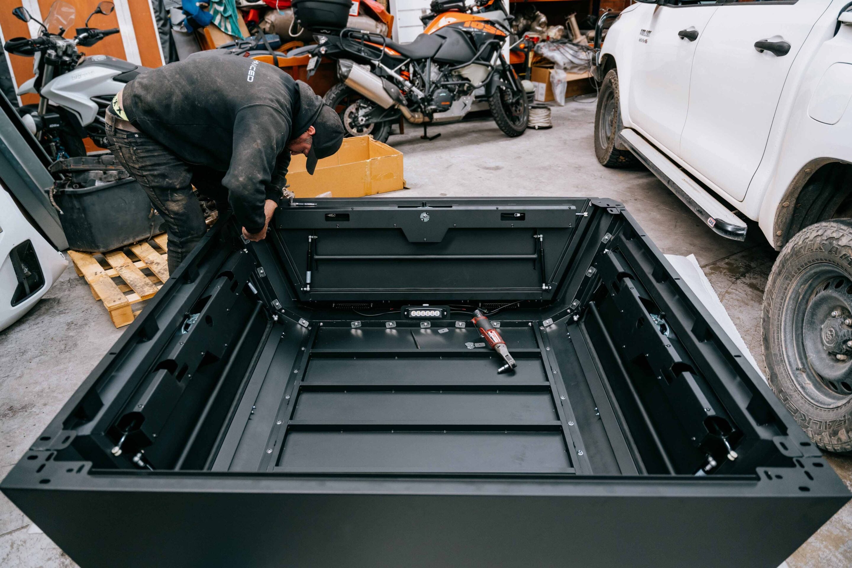 Worker in a black hoodie crouches over a large black metal frame, assembling it in a cluttered workshop with motorcycles and a white pickup in the background.