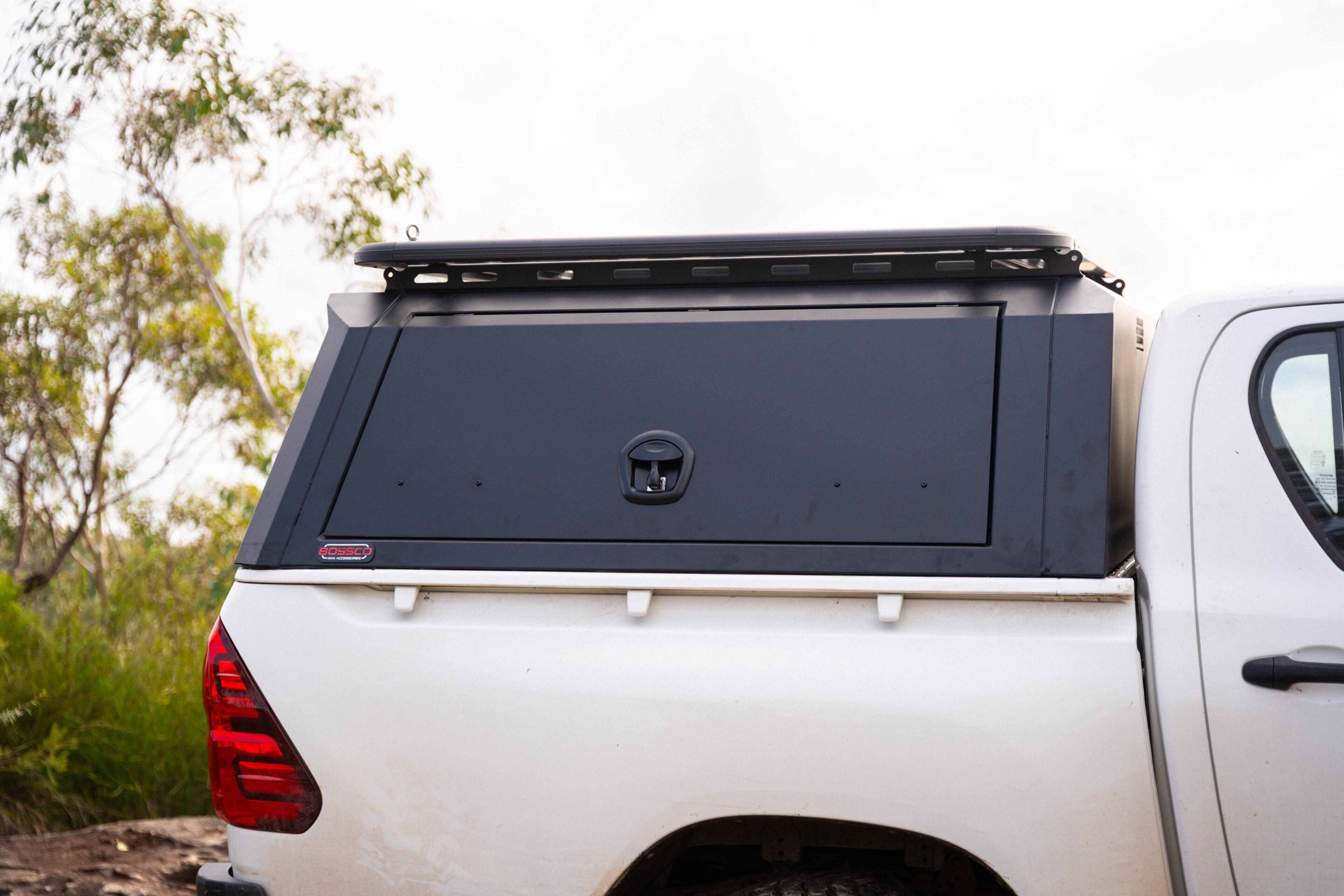 Rear view of a white pickup truck with a dark cargo canopy and roof rack, showing the tail light on the left and rugged hardware on top.