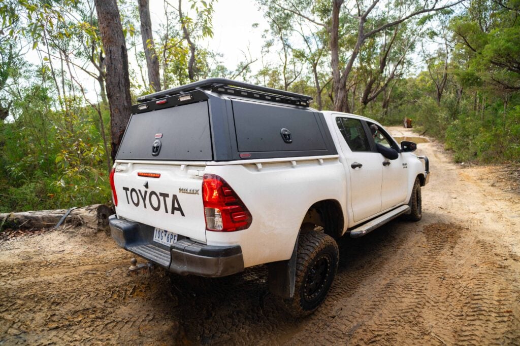 White Toyota pickup with a black camper shell parked on a dirt forest road. pocket-friendly camp gear on top rack visible in a wooded area.