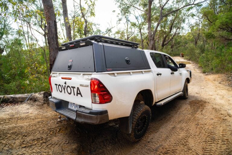 White Toyota pickup with a black camper shell parked on a dirt forest road. pocket-friendly camp gear on top rack visible in a wooded area.