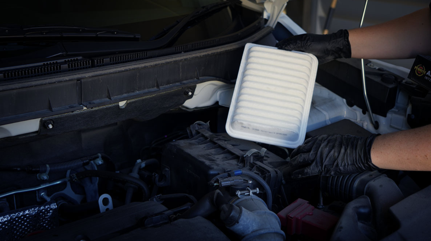 Technician wearing black gloves removes a white pleated car air filter from the engine bay under the hood.