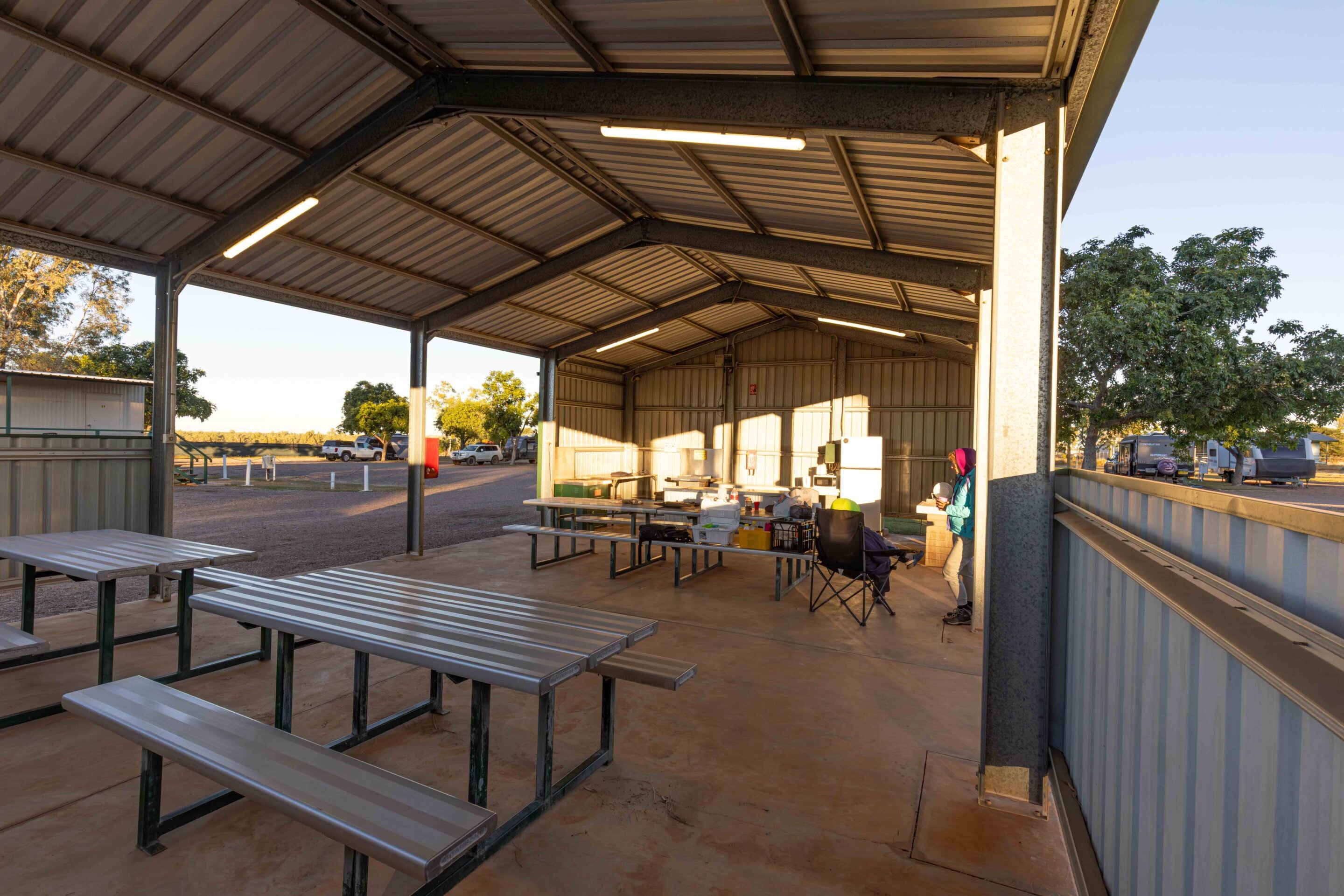 Open-air metal pavilion with picnic tables and stored equipment; sunlit exterior with trees and parked vehicles beyond.