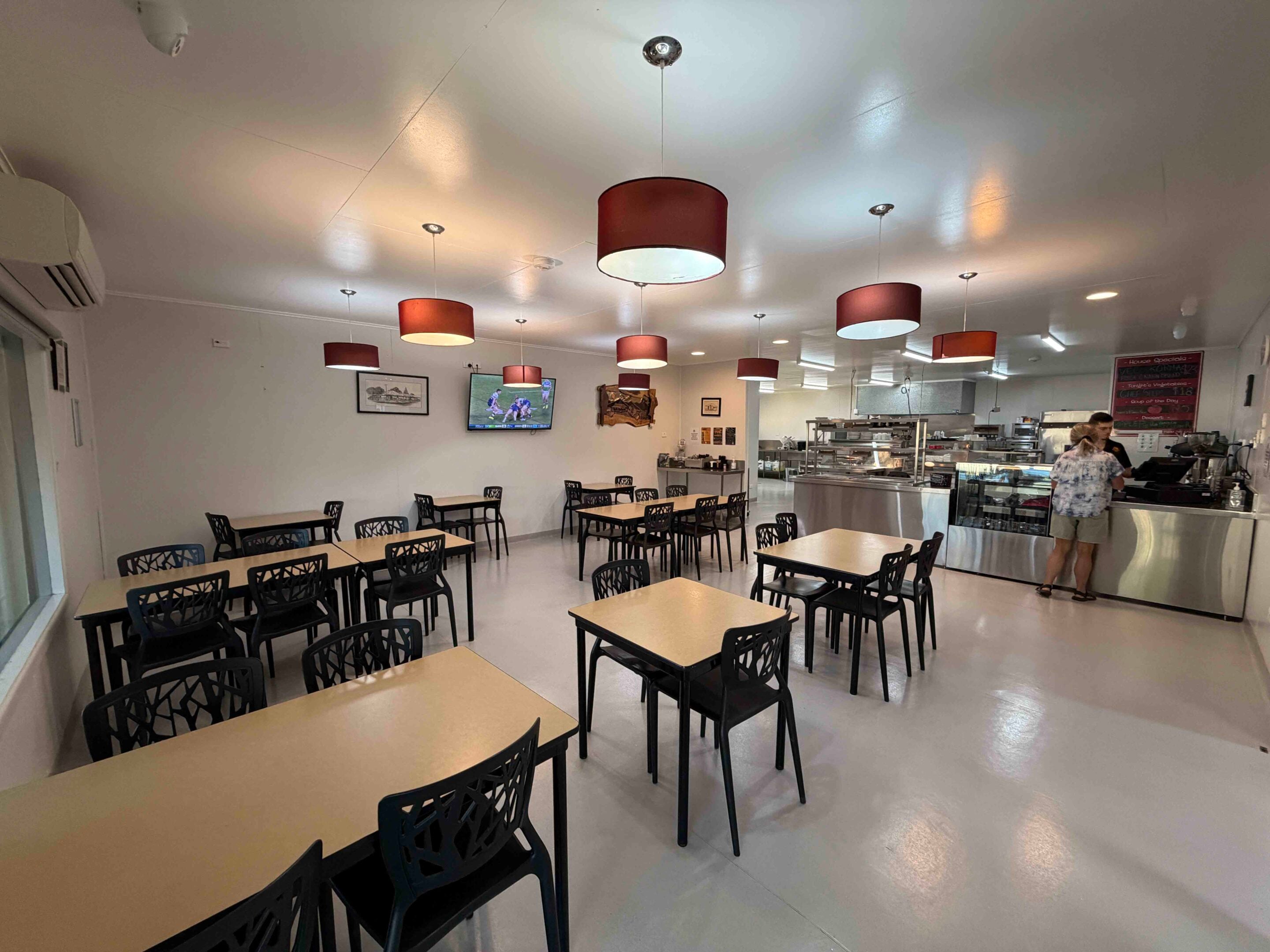Cafeteria with rows of light brown tables and black chairs, red drum pendant lights, and a service counter in the back.