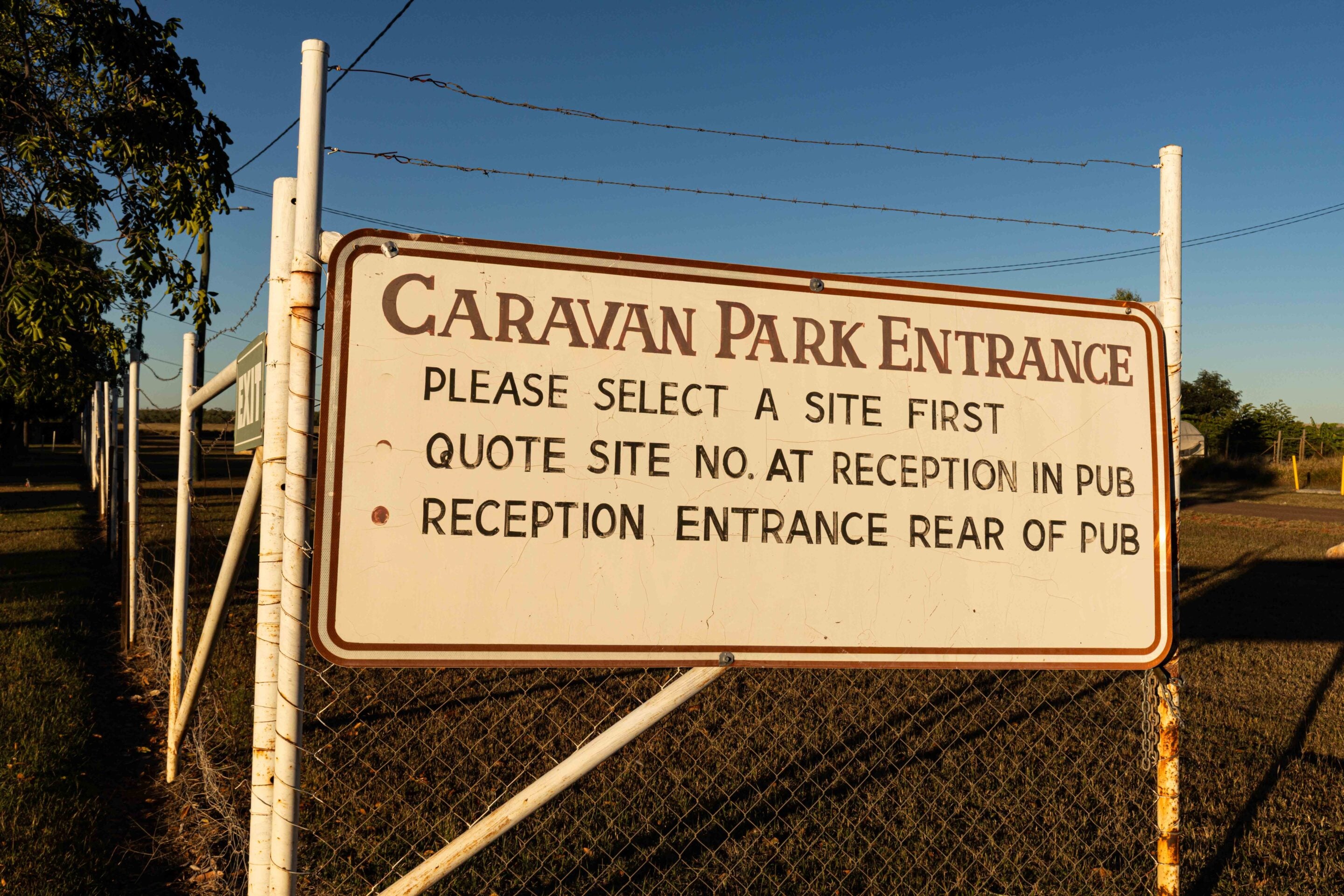 Large weathered sign reading Caravan Park Entrance mounted on a chain-link fence with barbed wire, outdoors under a clear blue sky.