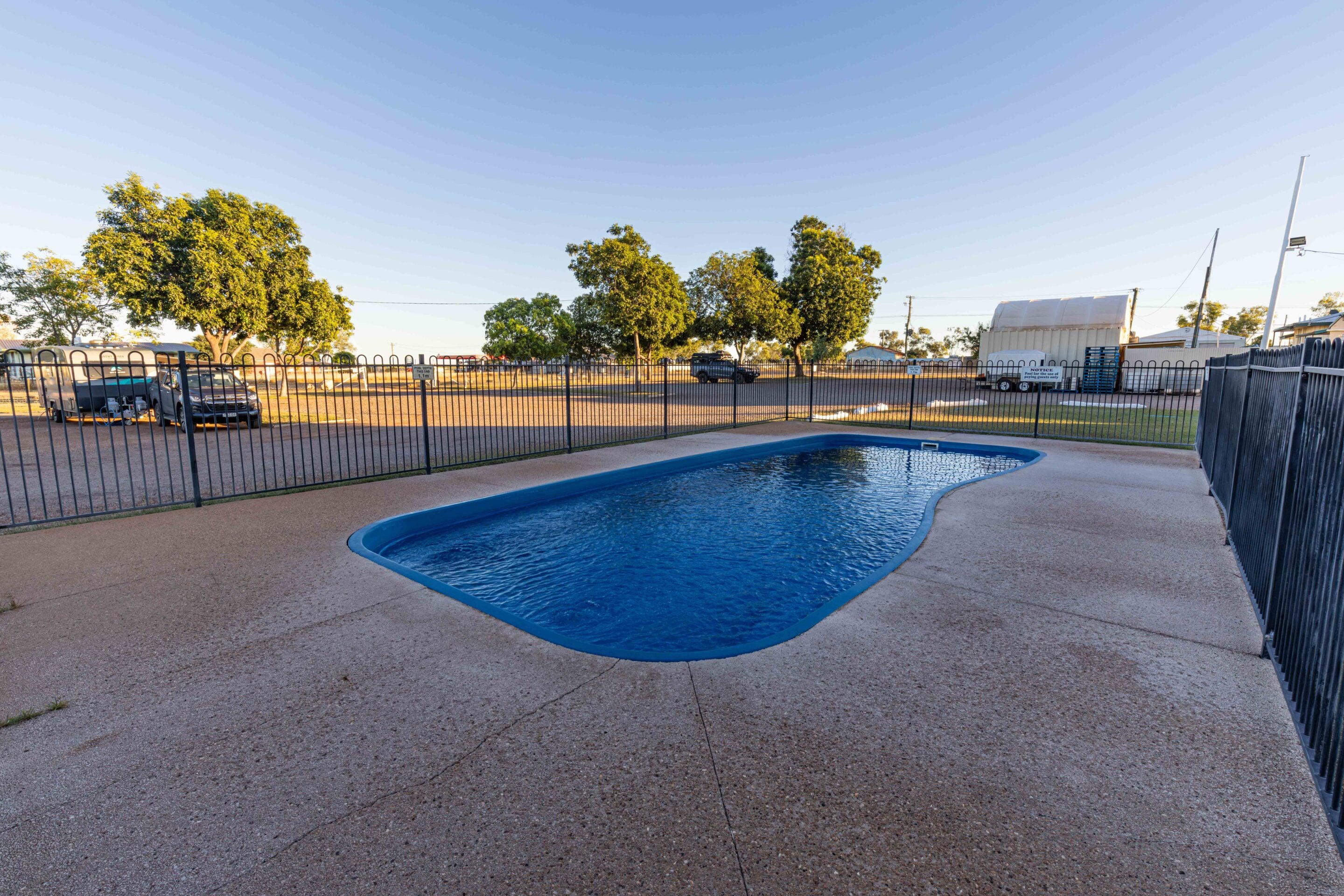 Curved blue pool surrounded by a black metal fence, with trees, parked vehicles, and a white trailer beyond in a sunny yard scene.