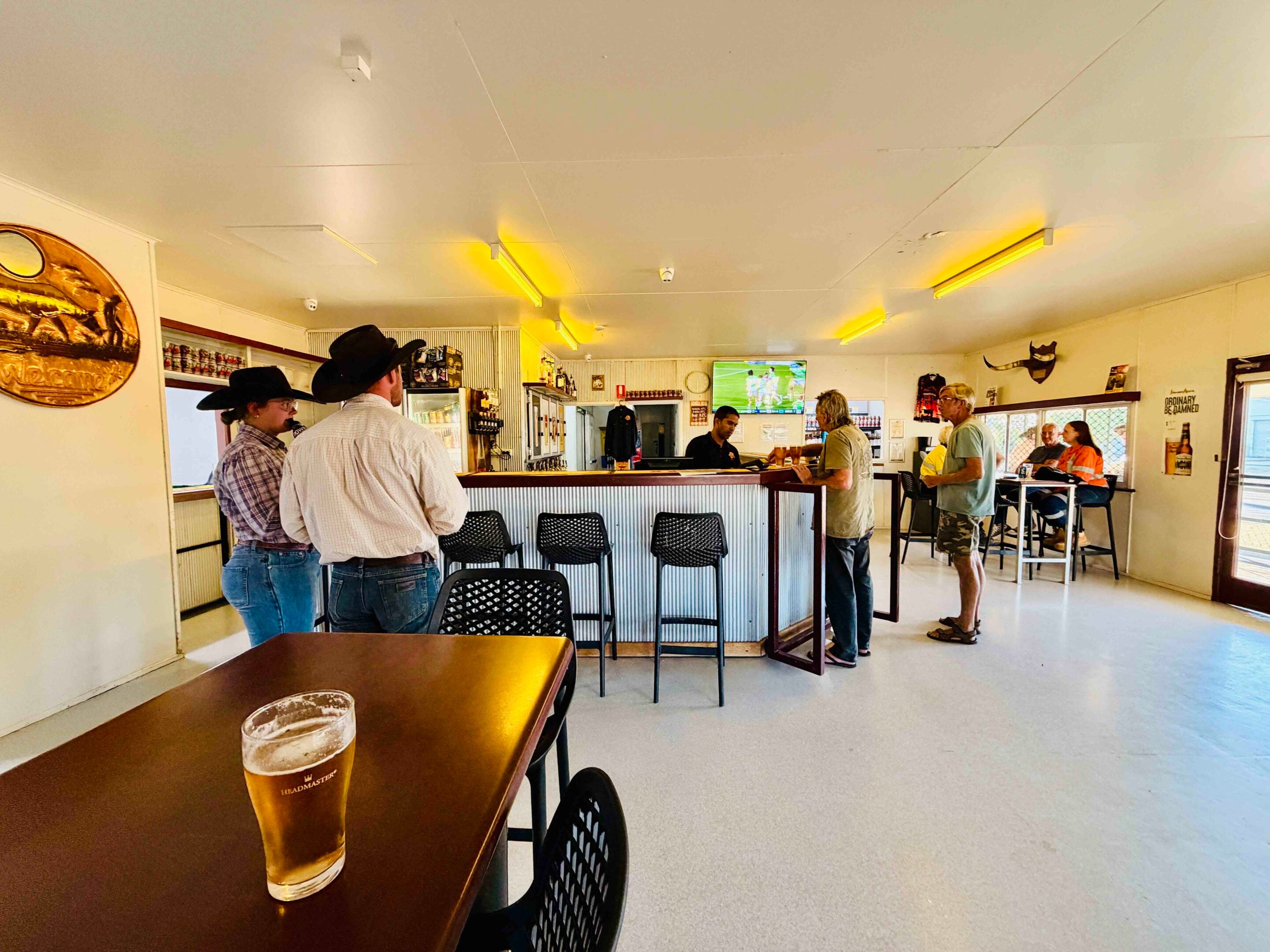 Inside a casual bar with patrons gathered at a high counter; a pint of beer sits on the foreground table.