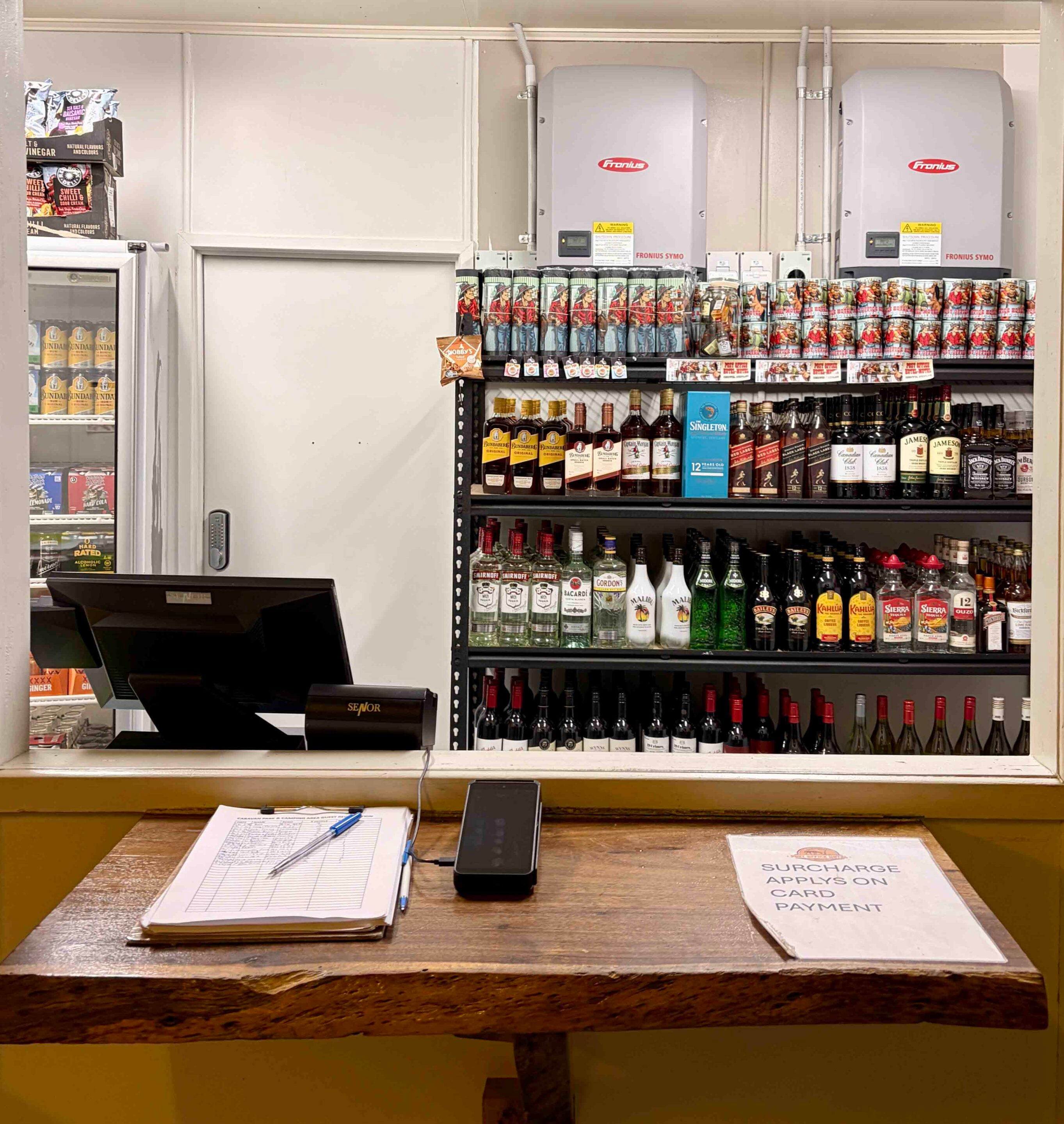Checkout counter in a liquor store with bottles on shelves behind, and two large gray Fronius machines mounted on the wall above the display.