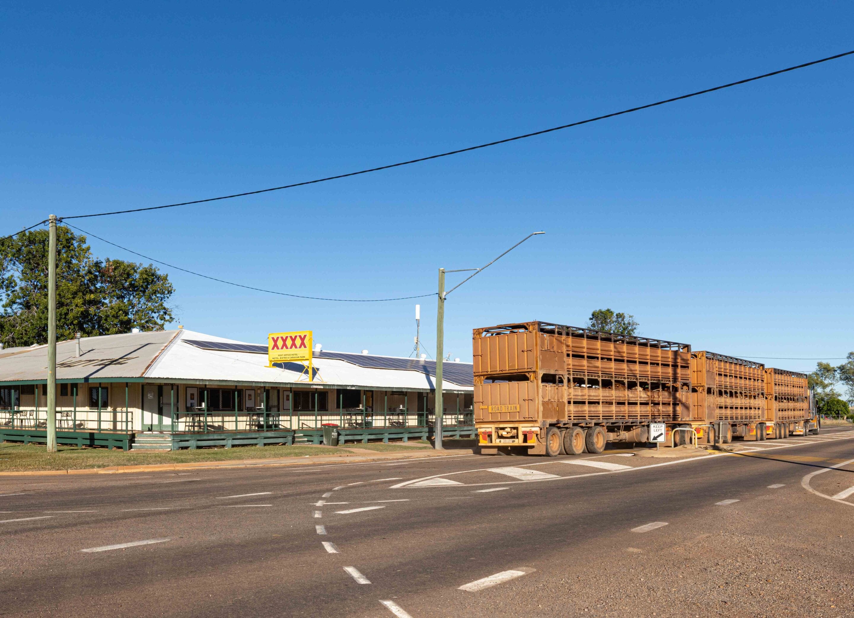 Wide street scene showing utility poles and power lines over a low building with a yellow sign, and a large brown pallet truck trailer loaded with stacked pallets against a clear blue sky.