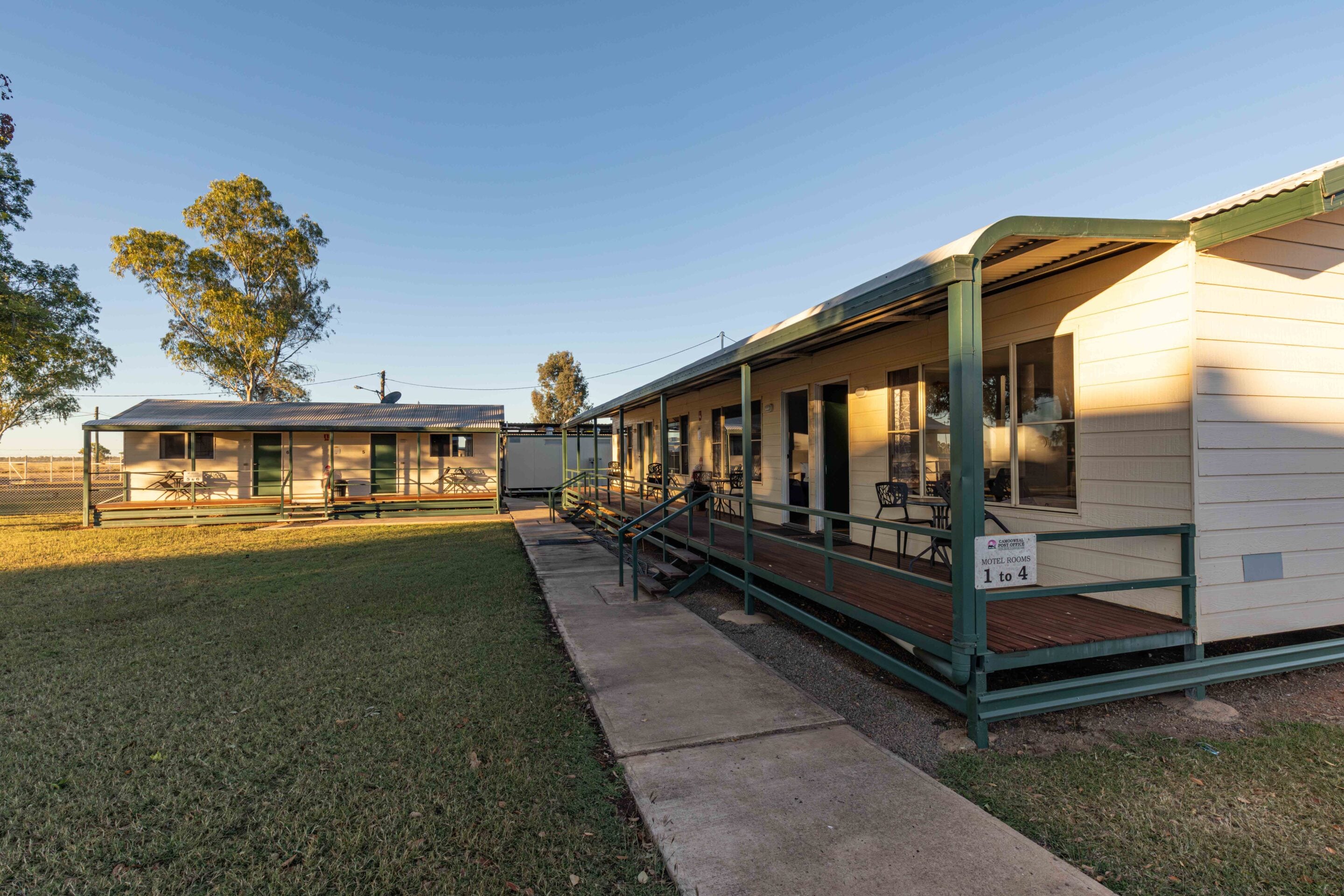 Wide view of a small motel with a covered walkway, green railings, and cream siding under a clear blue sky, trees in the background.