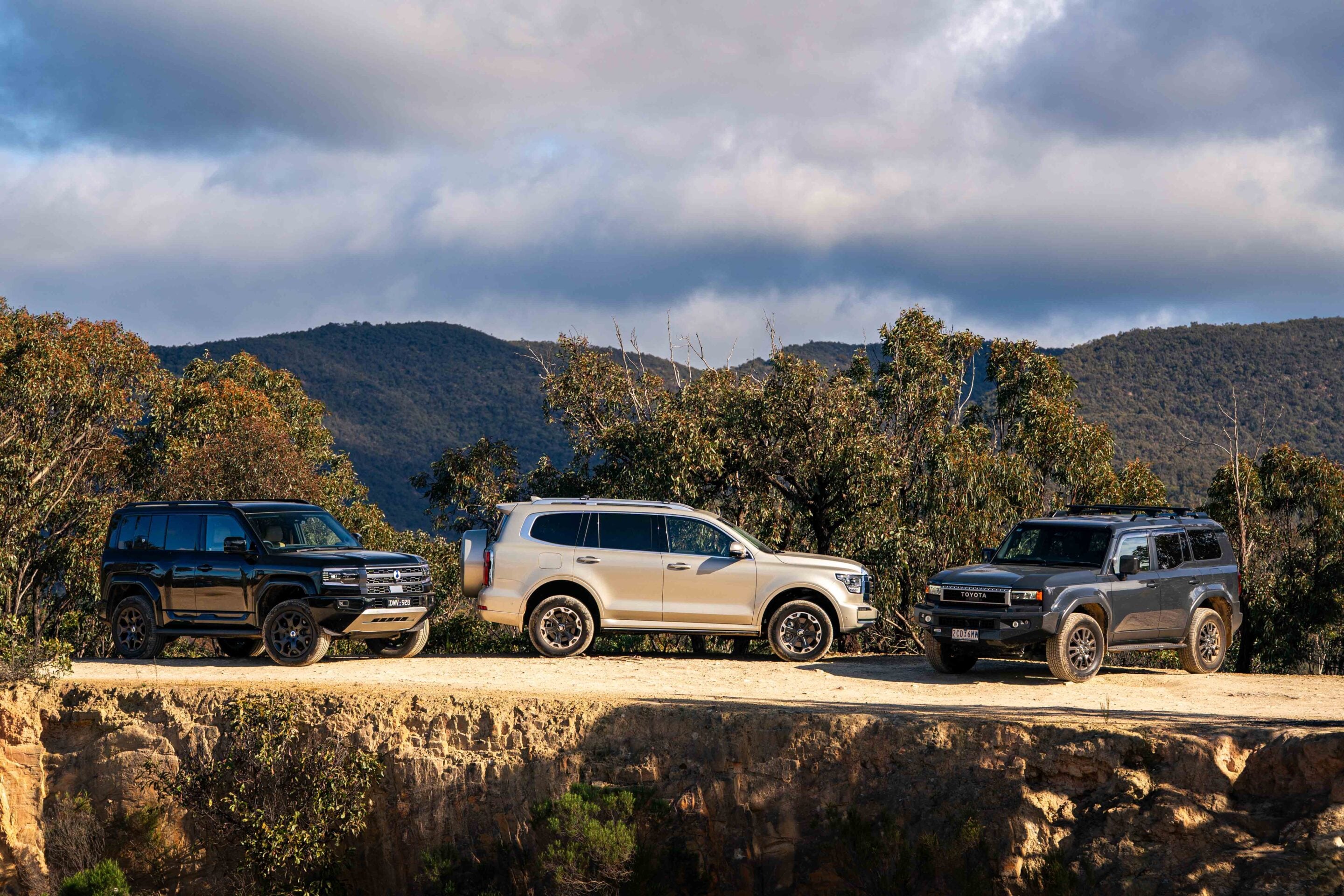 Line of four SUVs parked on a dirt lot at a canyon overlook with hills and a cloudy sky behind them.