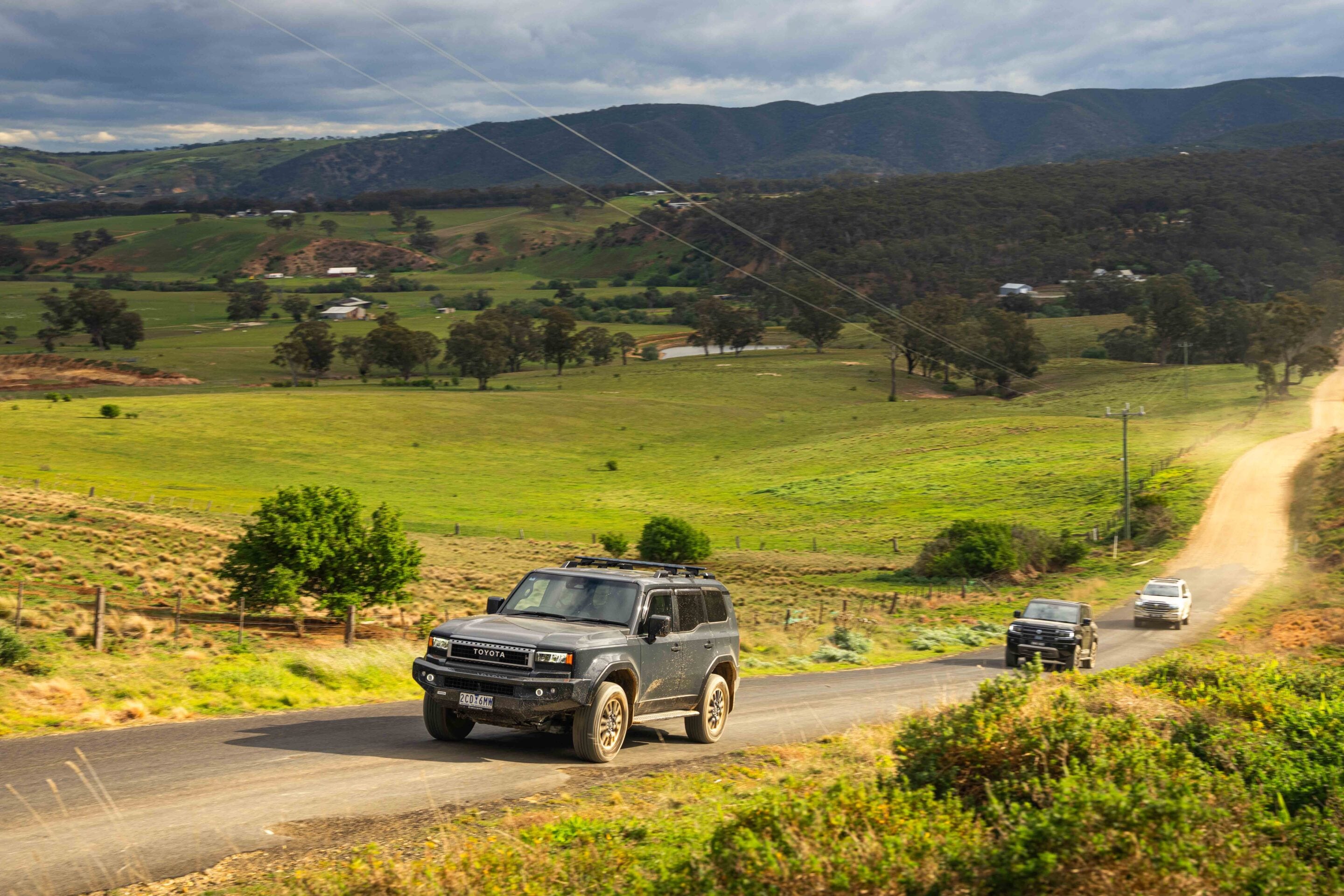 SUVs driving on a winding rural road through green fields and distant hills under a cloudy sky. Approaching traffic with a Toyota SUV in the foreground.