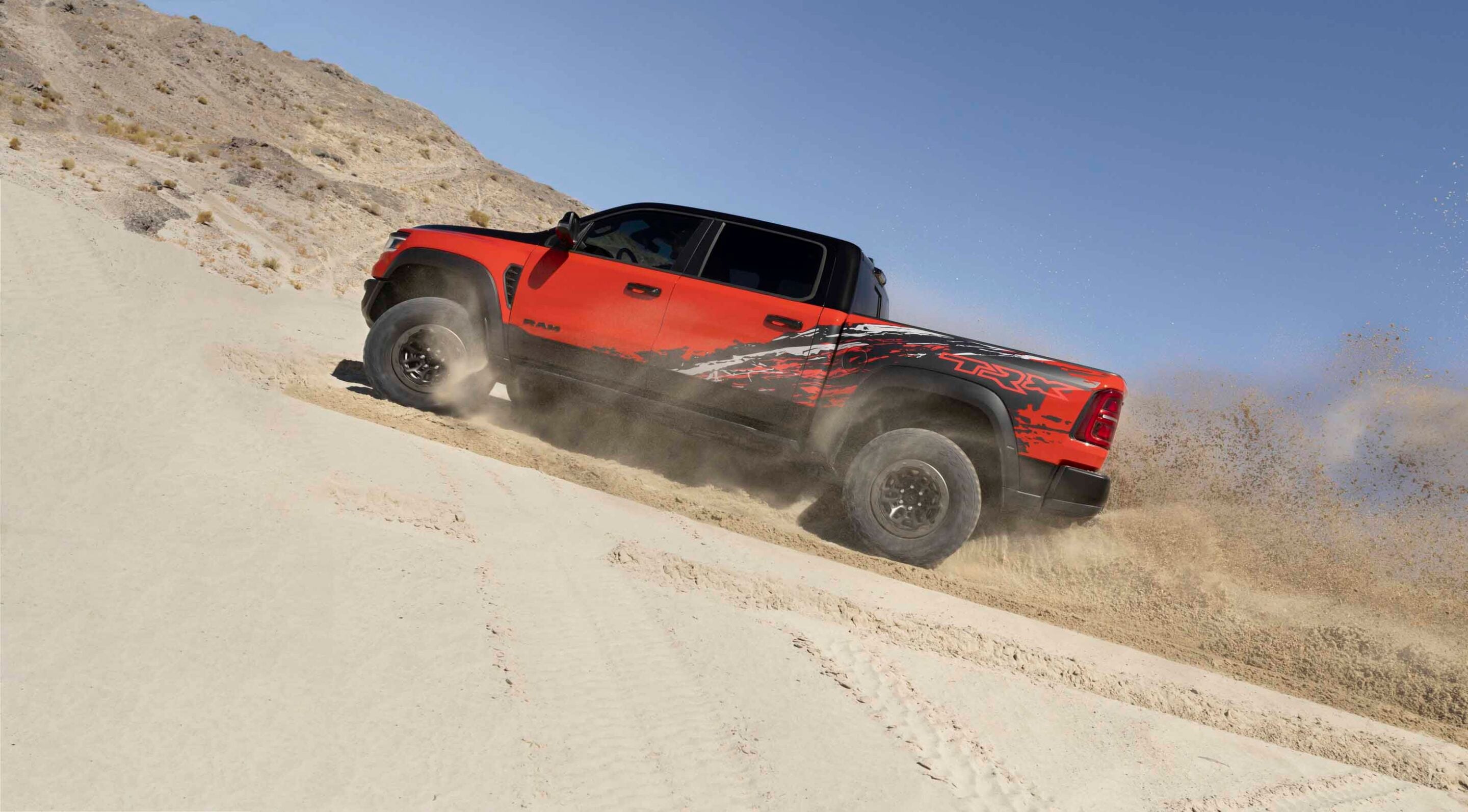 Red and black off-road pickup ascending a sandy desert dune, kicking up dust under a clear blue sky