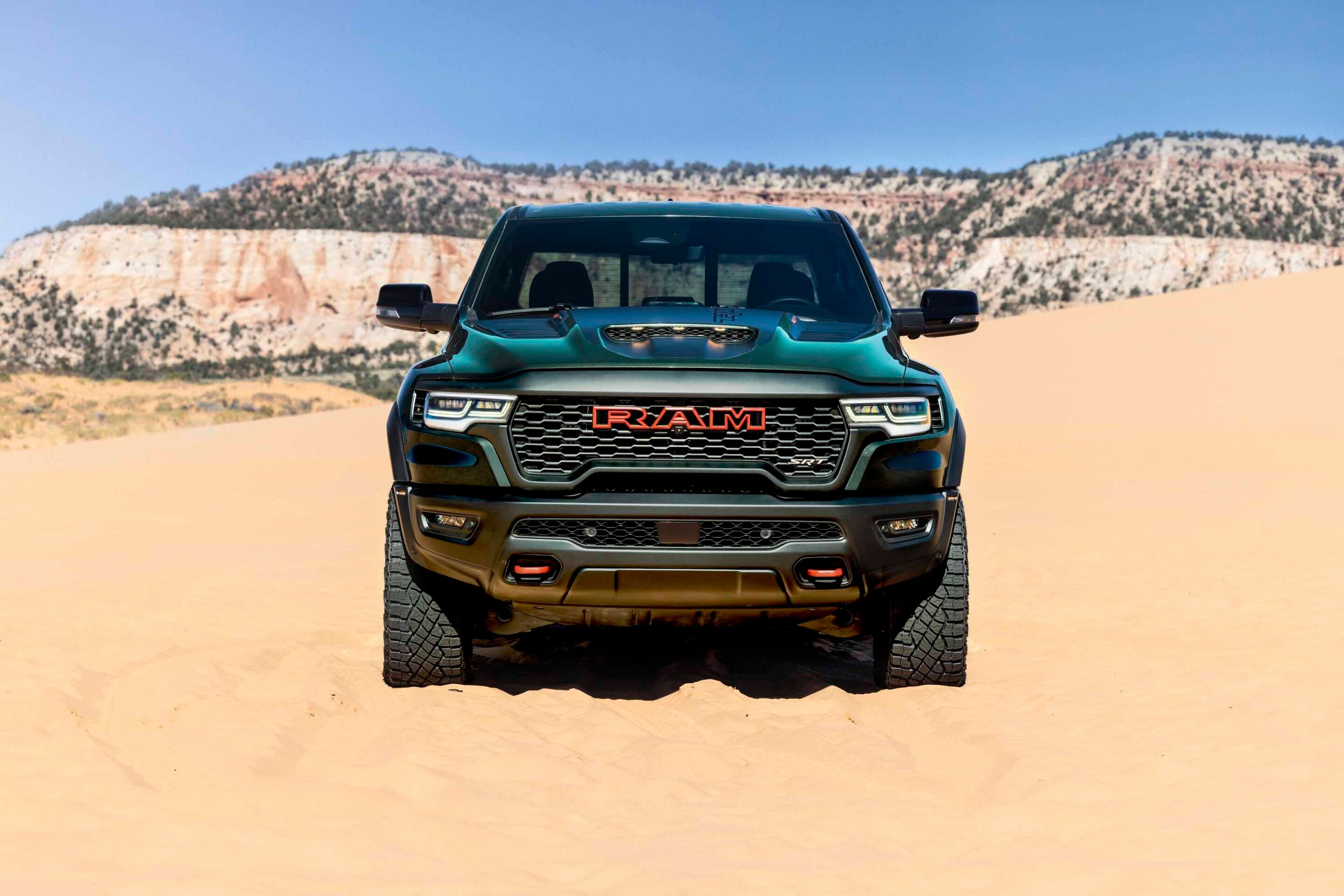 Front view of a dark teal RAM pickup truck parked on a sandy desert with rocky hills in the background.