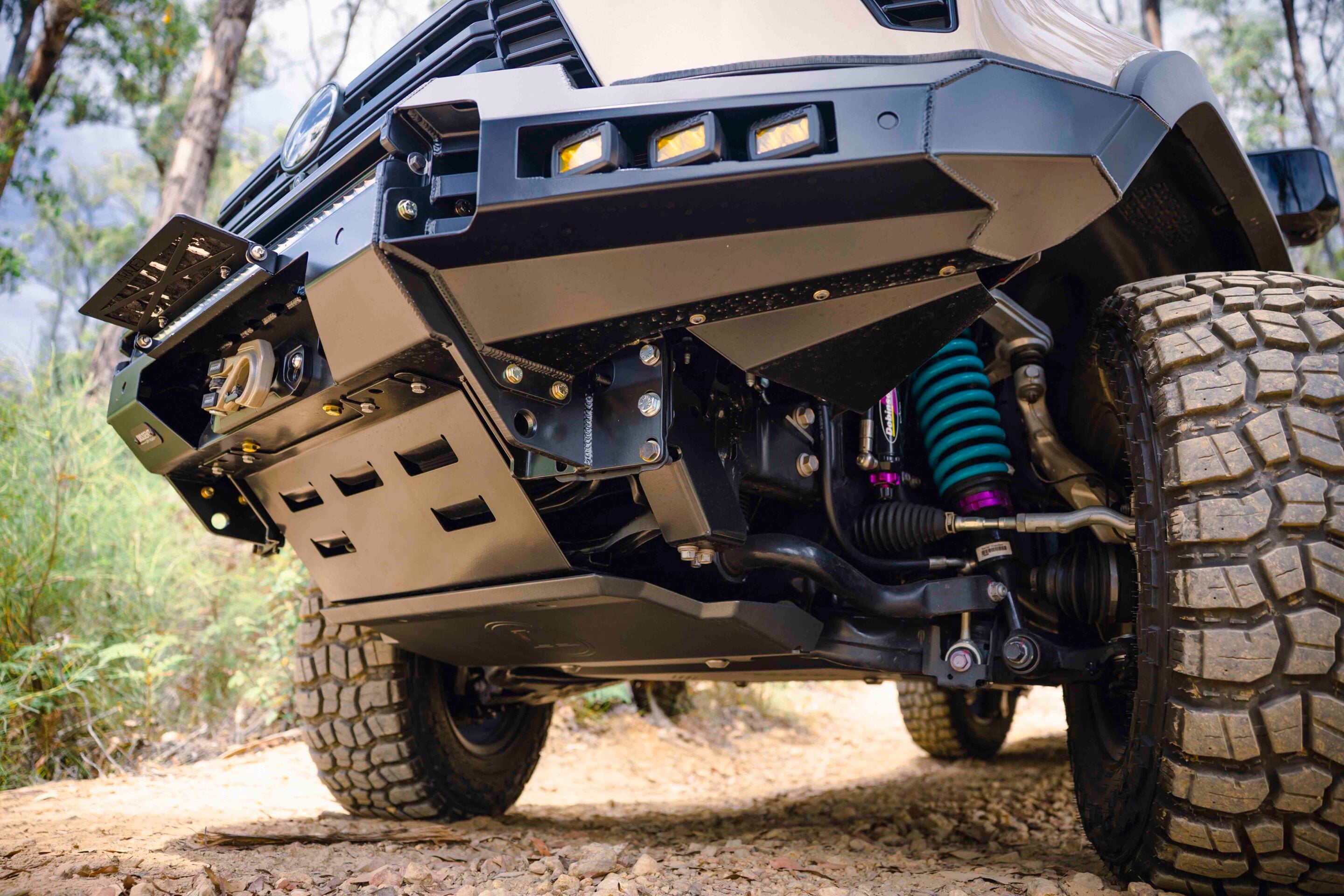 Close-up of a heavily modified off-road truck's front bumper, winch, and rugged tires on a dirt trail.