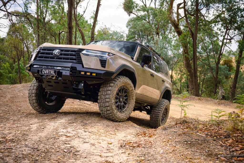 Beige off-road SUV angled on a dirt trail in a wooded area, large tires and roof rack visible.