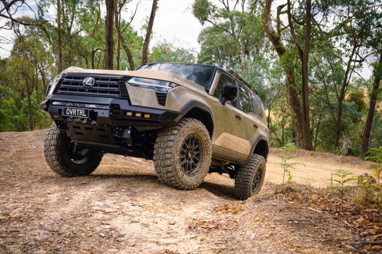 Beige off-road SUV angled on a dirt trail in a wooded area, large tires and roof rack visible.