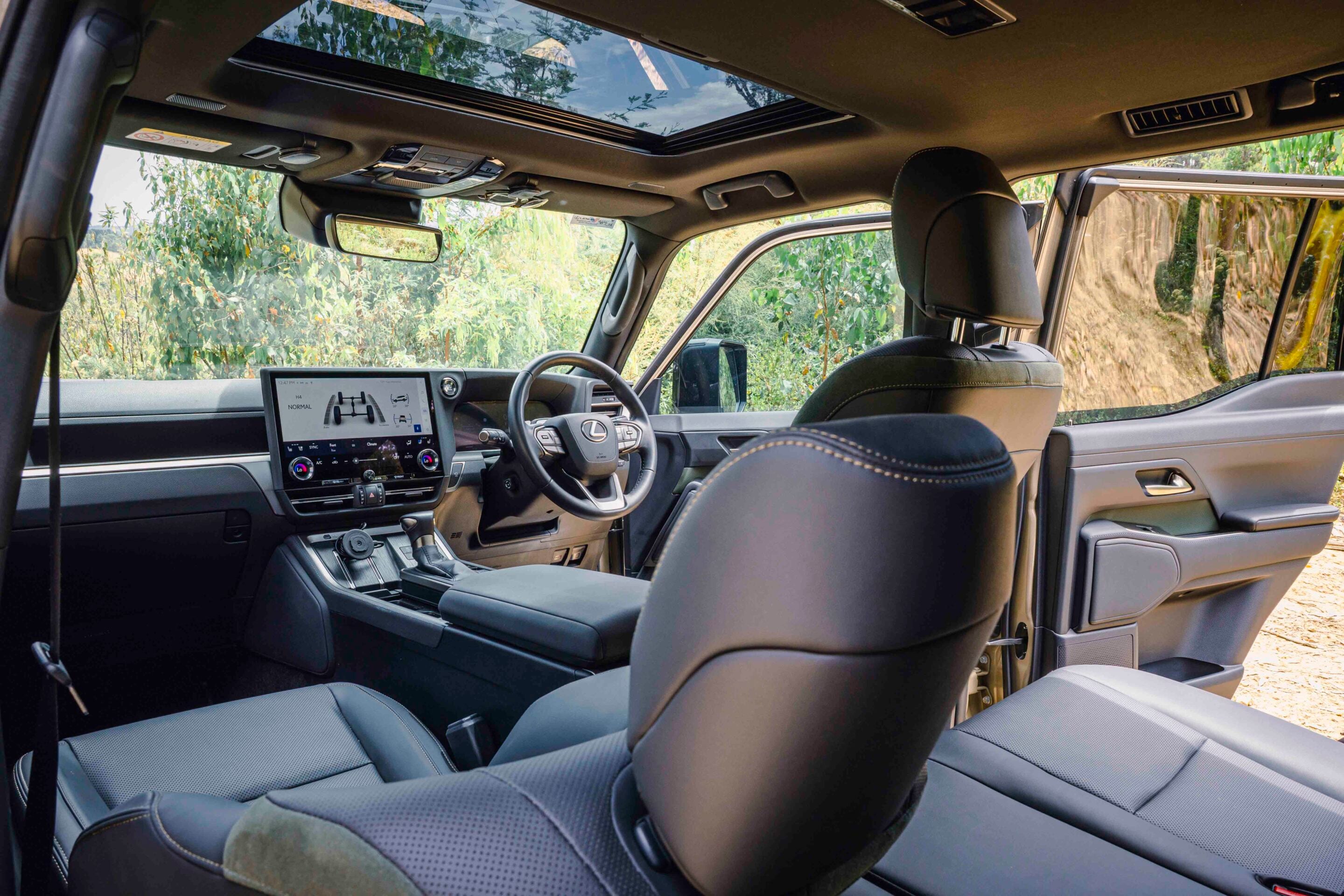 Front cabin of a modern SUV with a panoramic sunroof, large touchscreen, and black leather seats seen from the back seat perspective.