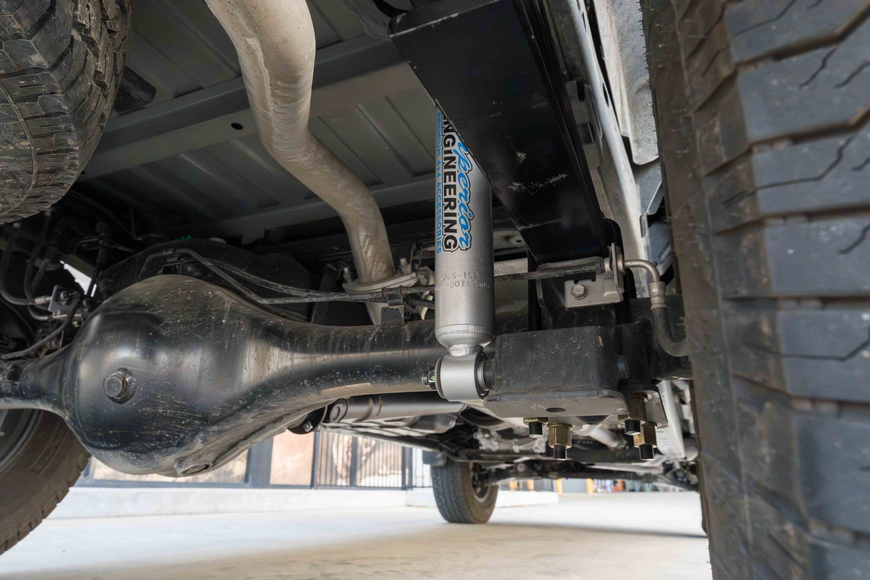 Underbody view of a vehicle showing the exhaust, suspension, and tire; a shock absorber has a blue 'Racing' sticker nearby