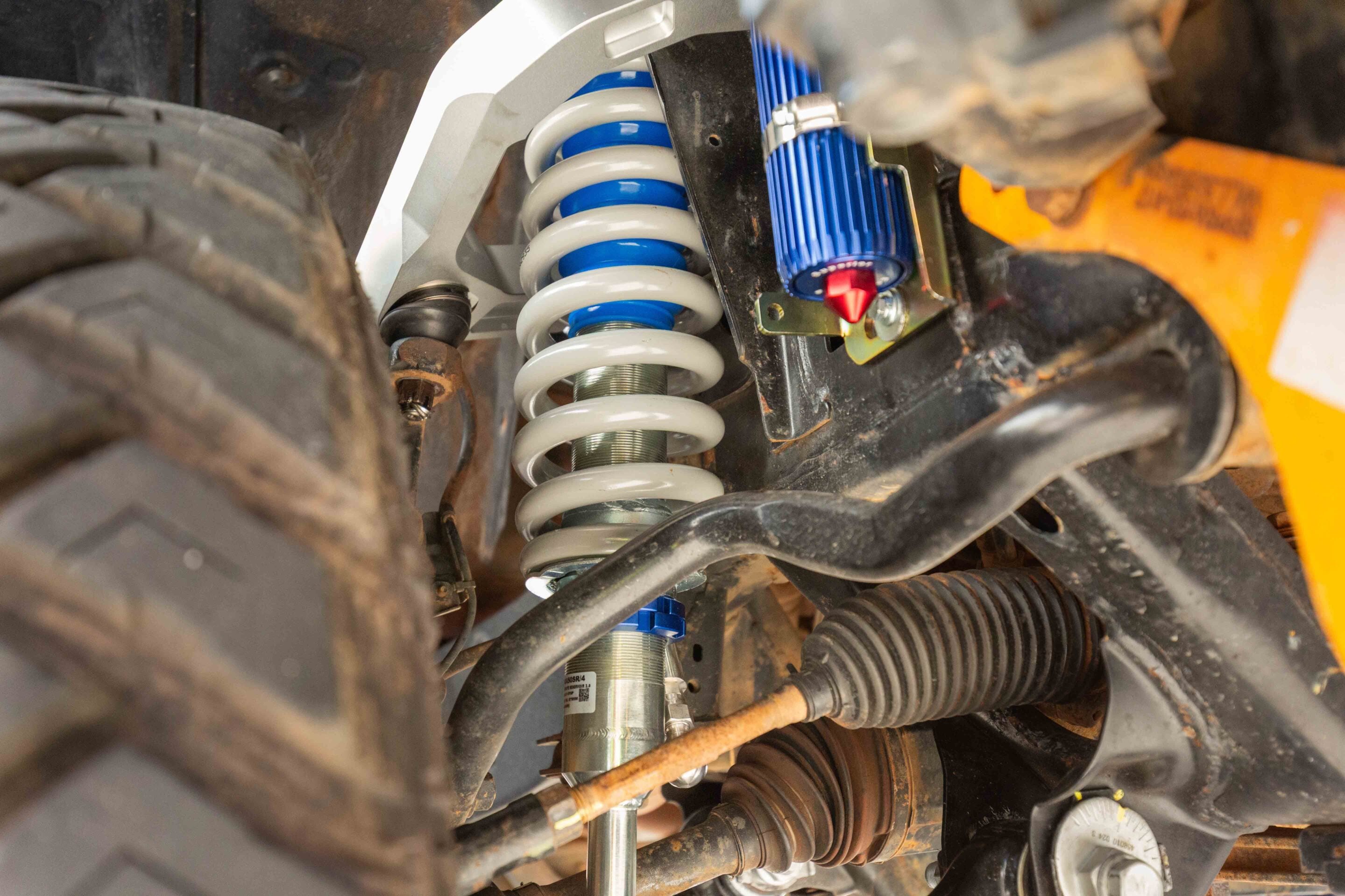 Close-up of a blue-and-white coil spring shock absorber in a vehicle suspension, with rusty and dusty surrounding parts.